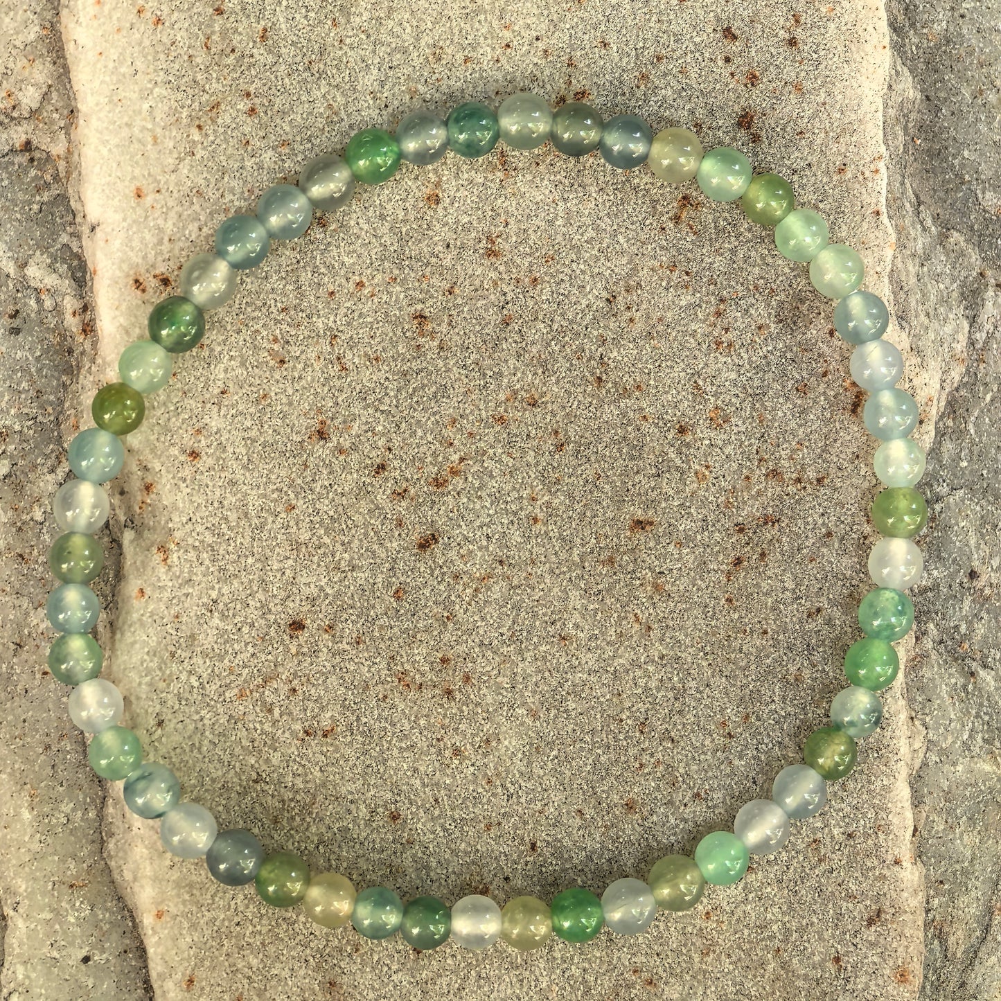 Natural green multicolor jadeite bracelet laid flat on stone background with 50 small round beads.
