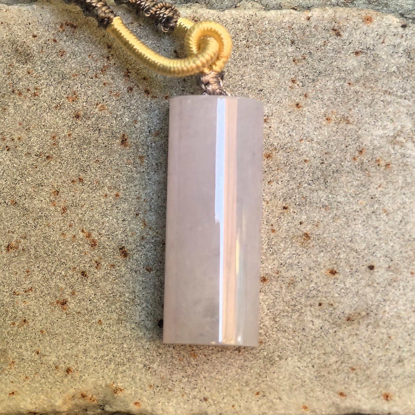 Light purple jadeite plain pillar pendant displayed on stone background with cord attached.
