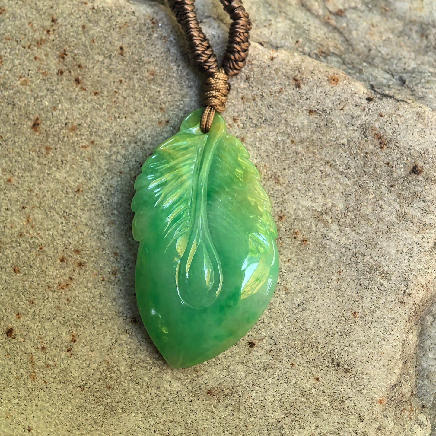 Glutinous green jadeite leaf pendant displayed on stone surface with natural light reflection.