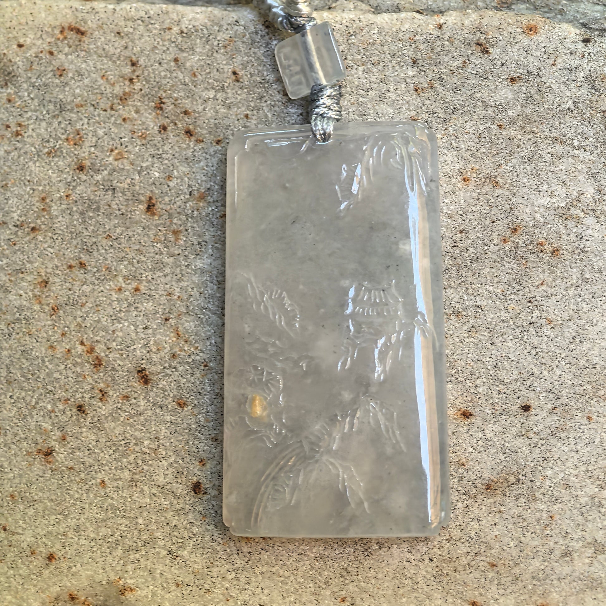 Rectangular white jadeite landscape pendant displayed on stone background with cord attached.