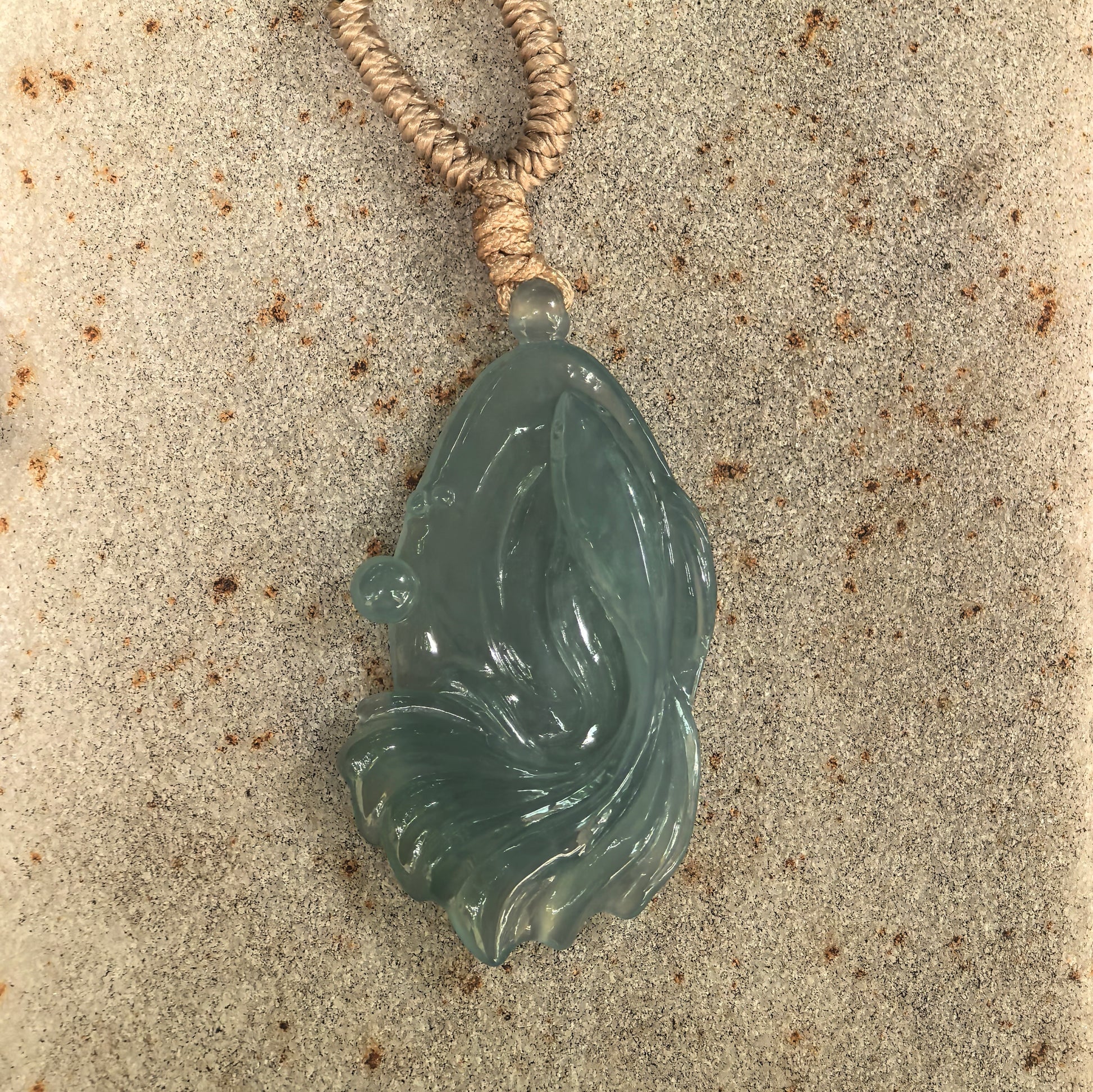 Close-up of deep green fish jadeite pendant on stone surface showing icy glutinous texture and goldfish in pond-inspired design.