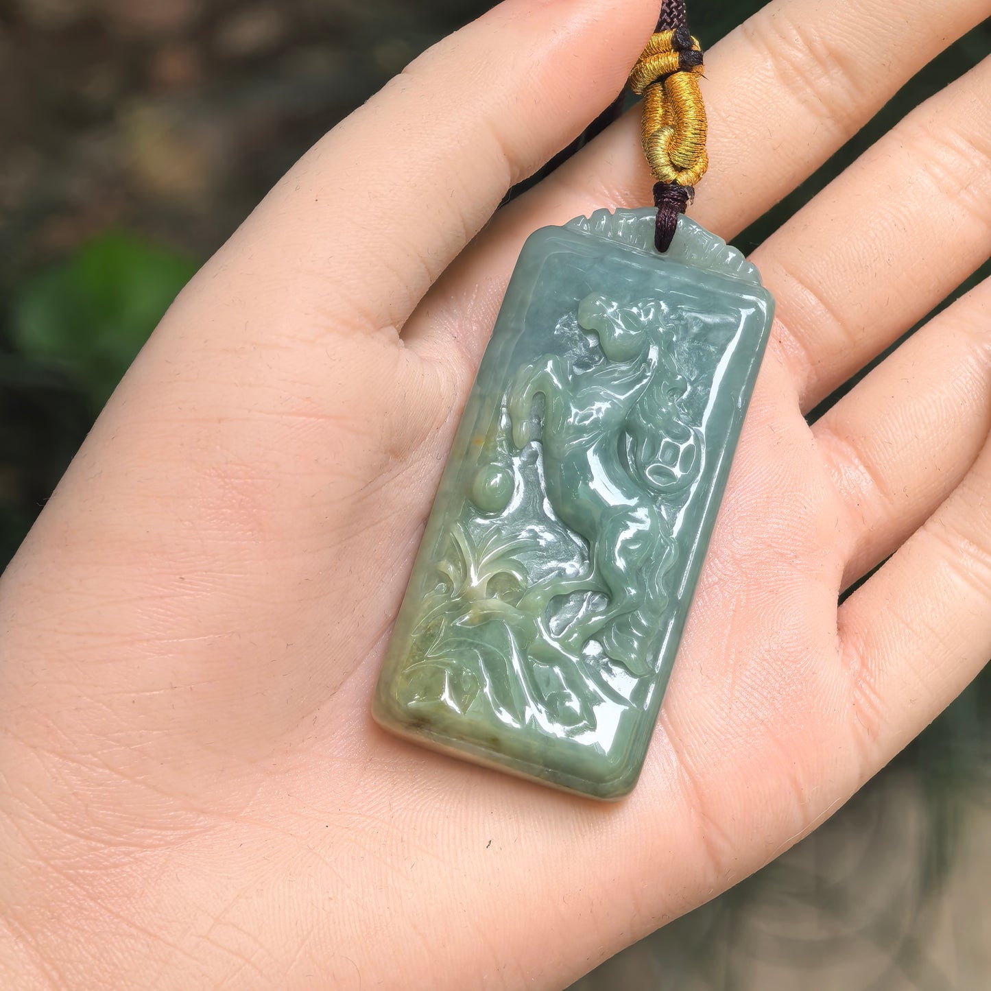 Deep green horse jadeite pendant shown in hand with raised horse carving and smooth polished surface.