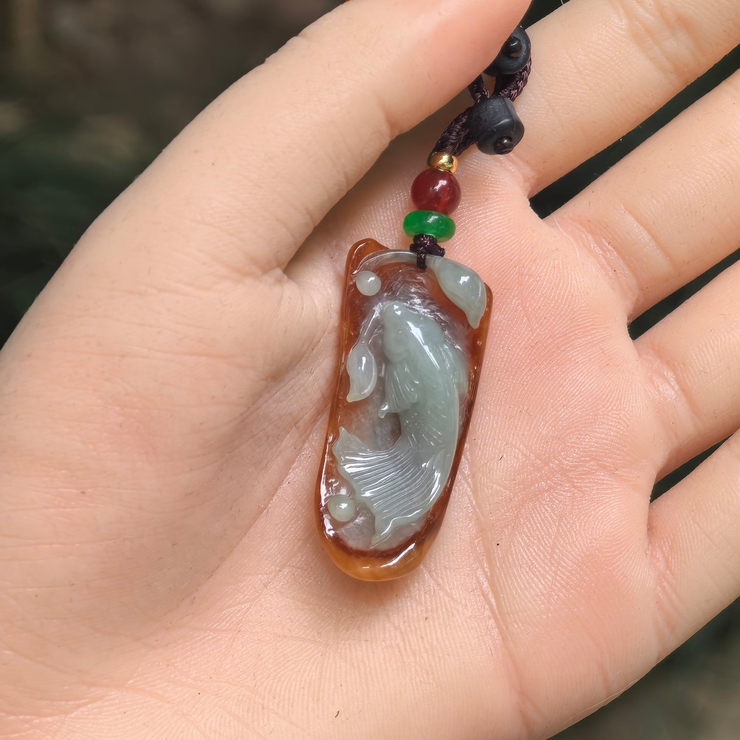 Carved fish jadeite pendant shown in hand with yellow-brown base and pale green fish motif.