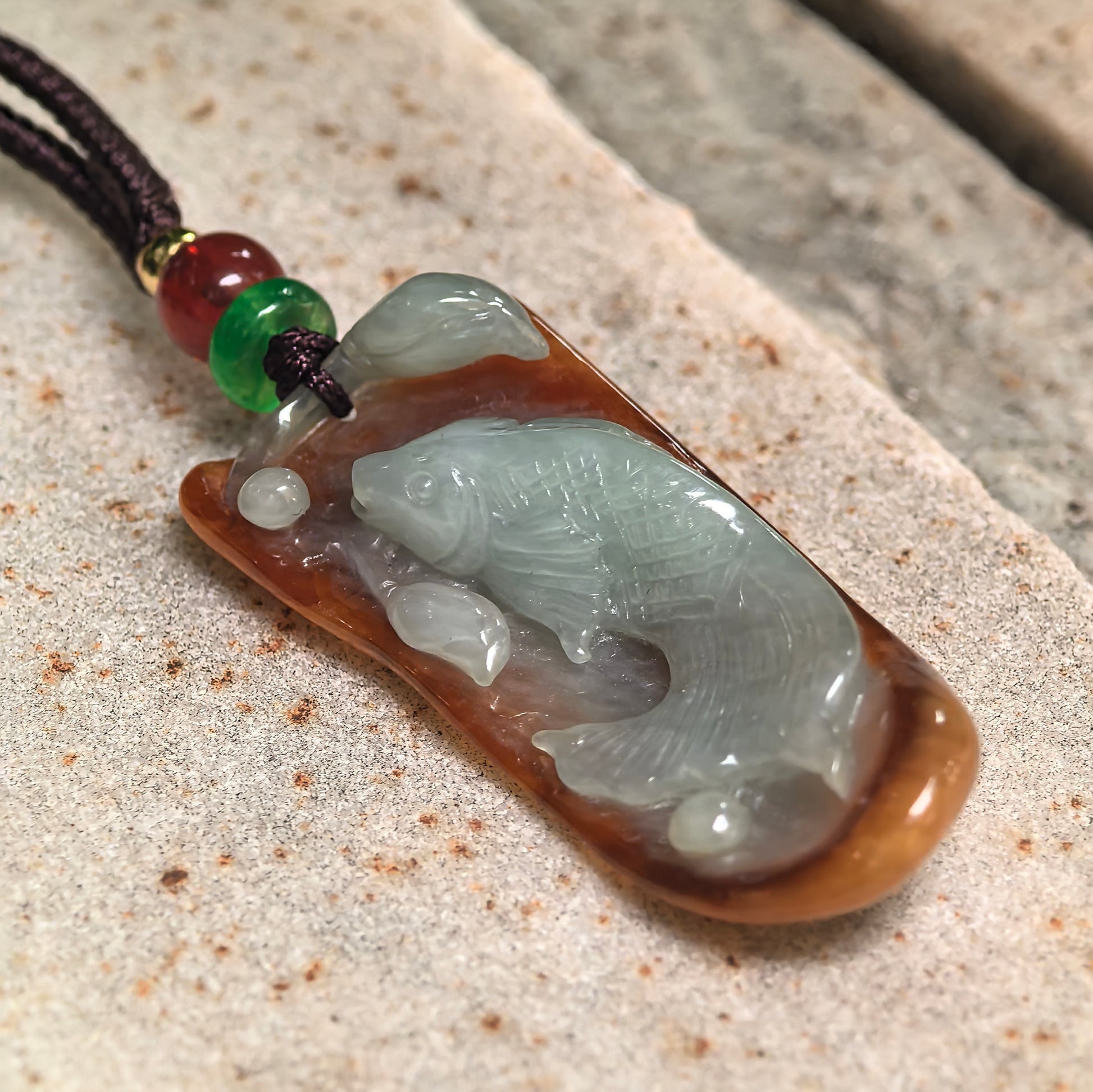 Angled view of yellow-brown jadeite pendant with pale green fish carving and rounded plaque shape.