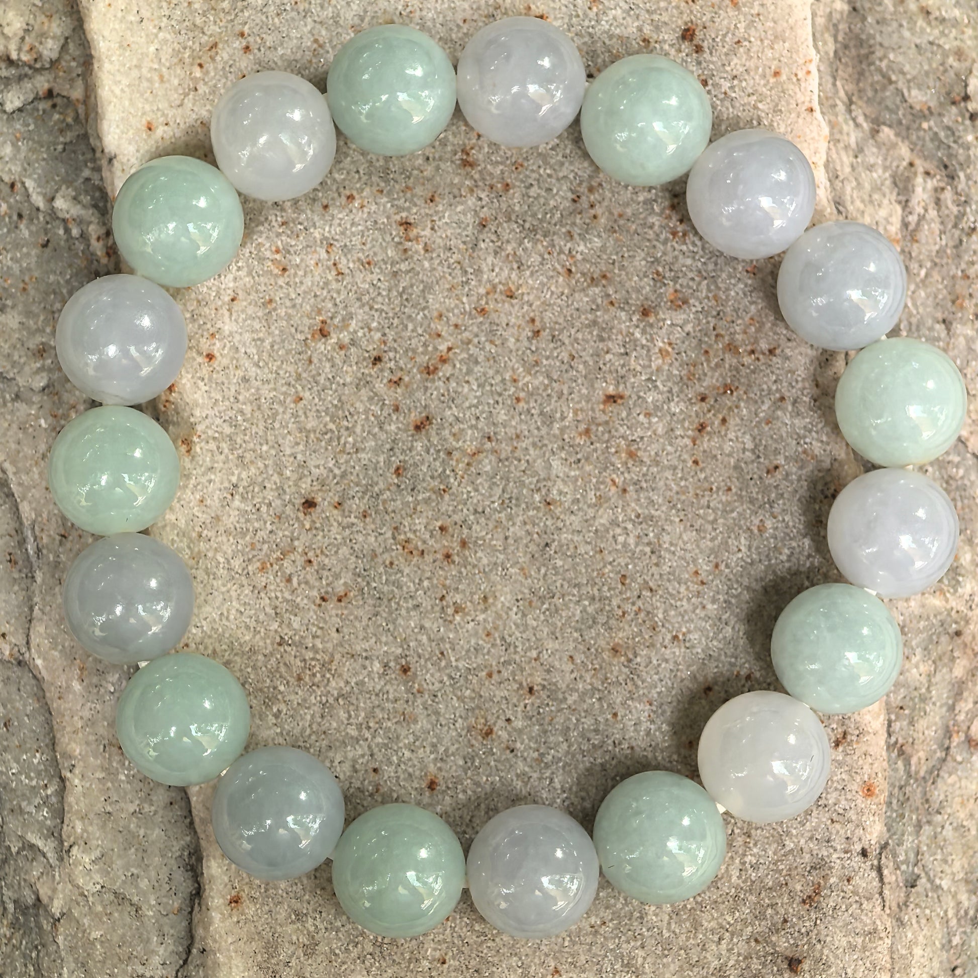 Pale green and lavender jadeite bead bracelet displayed on stone background.
