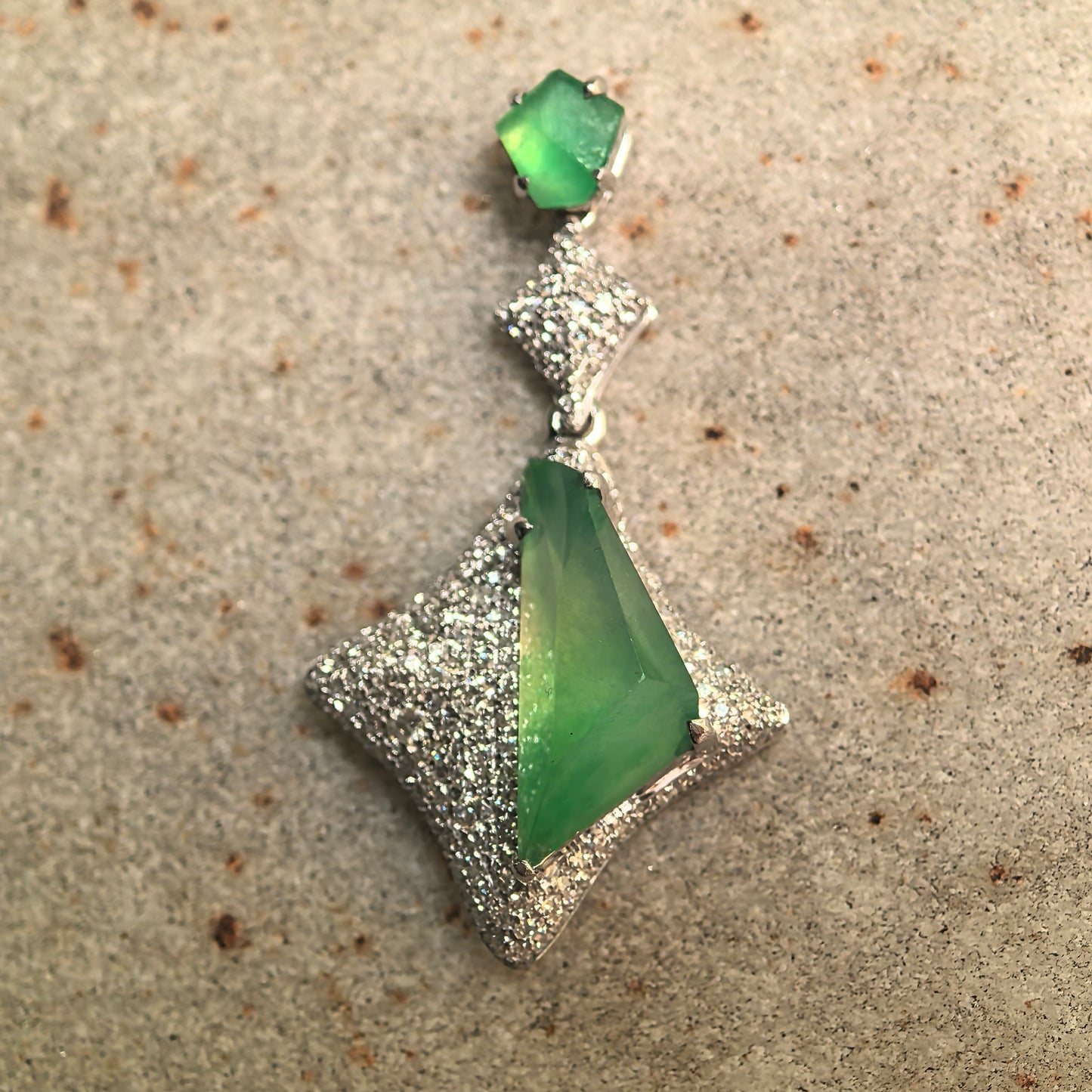 Green jadeite pendant with diamond-accented rhombus design displayed on stone surface.
