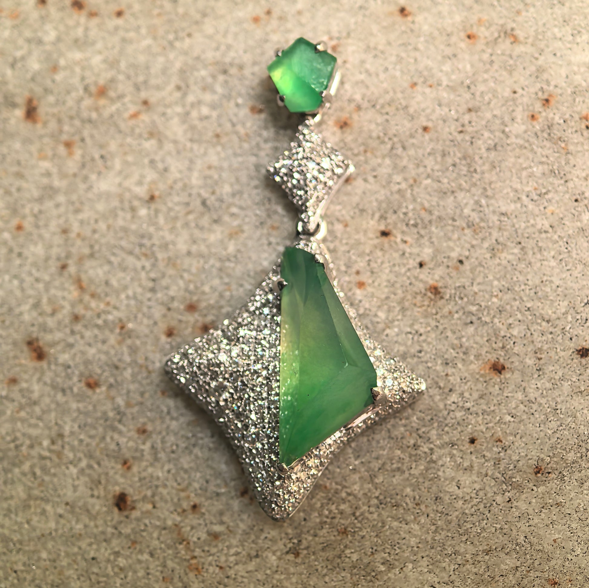 Green jadeite pendant with diamond-accented rhombus design displayed on stone surface.

