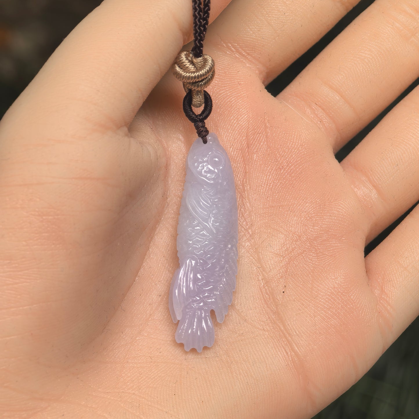 Three-dimensional carved jadeite fish pendant displayed on the hand with soft purple tone.
