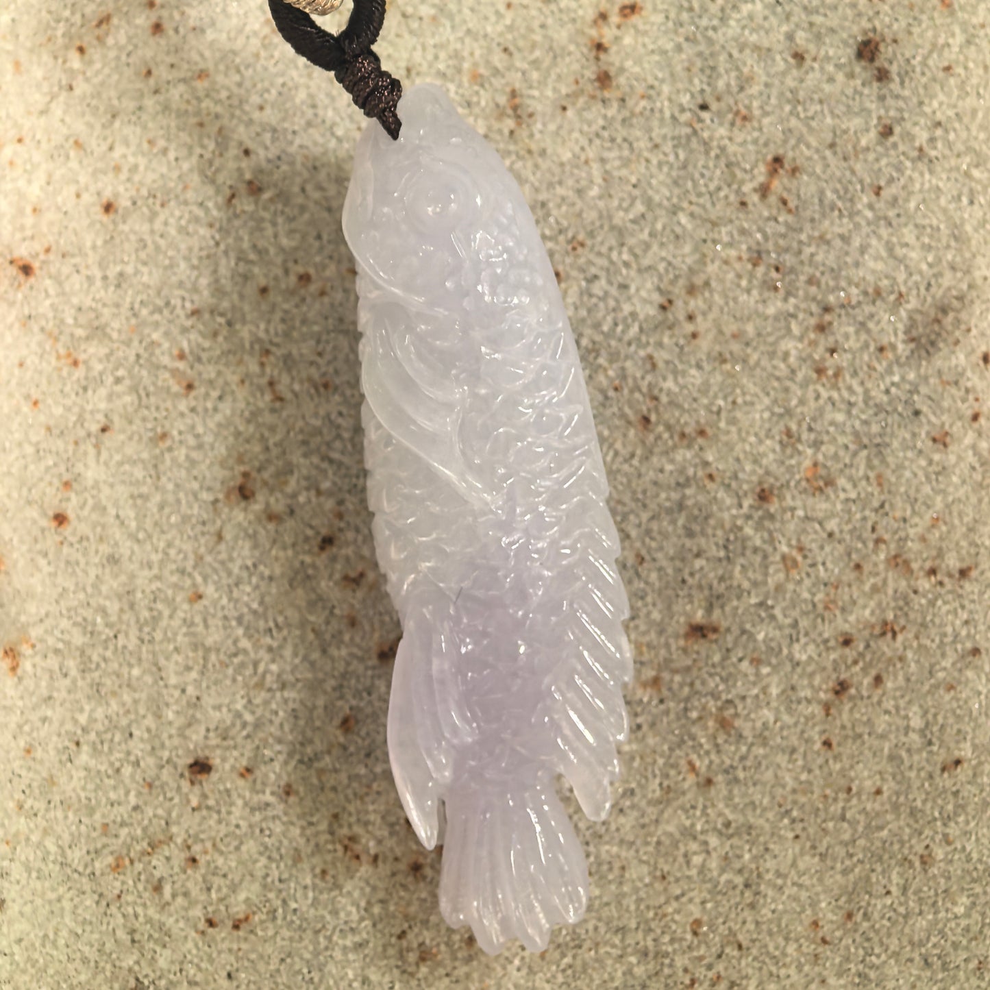 Close-up of a light lavender jadeite fish pendant showing carved scales and fins.
