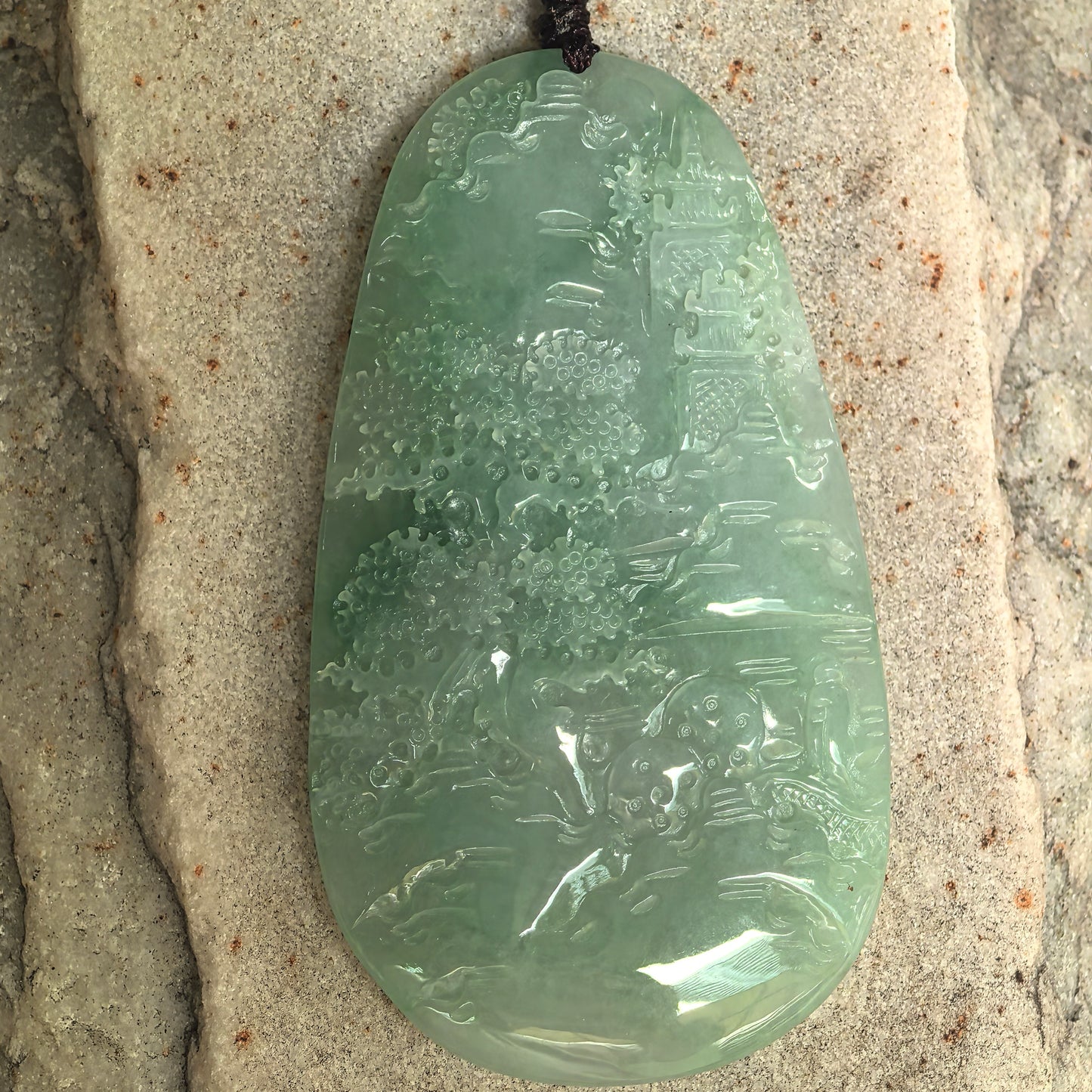 Carved jadeite landscape pendant showing trees, architecture, and flowing scenery.
