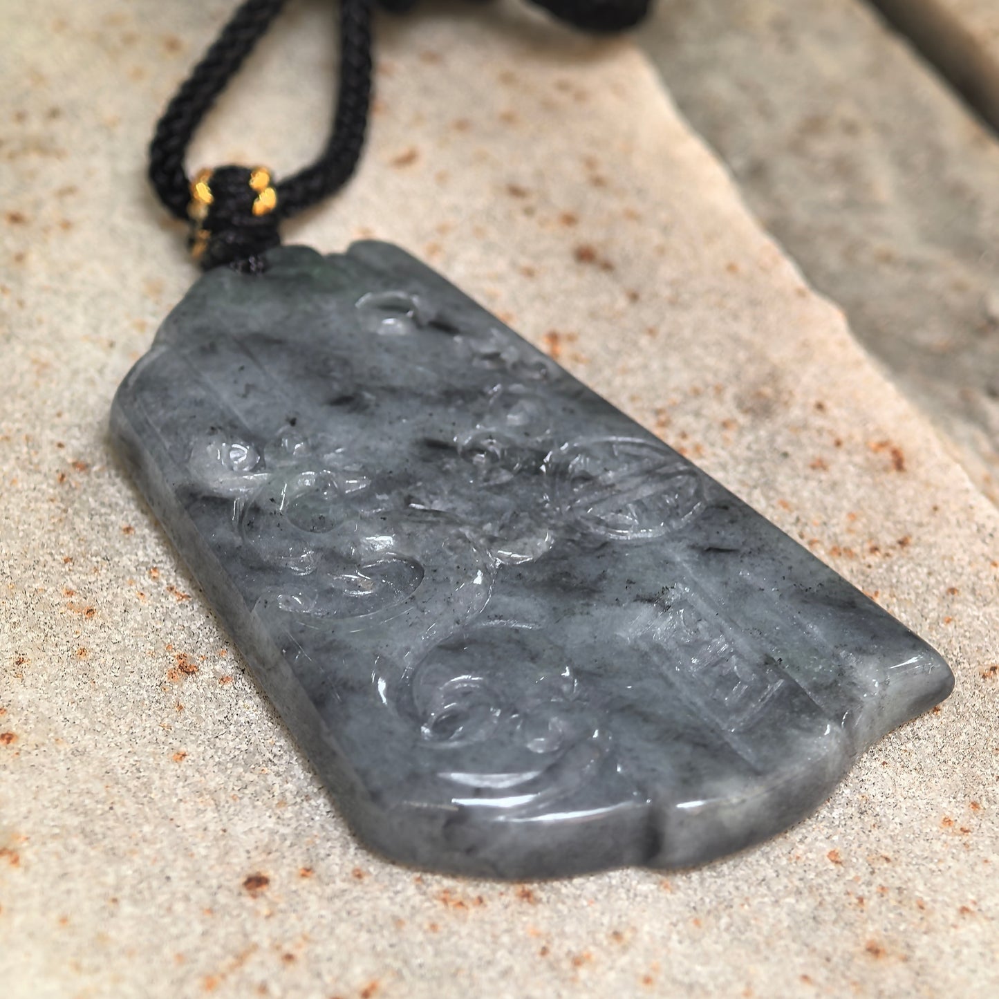 Side view of gray-black carved jadeite pendant showing its slim plaque form and glossy luster.
