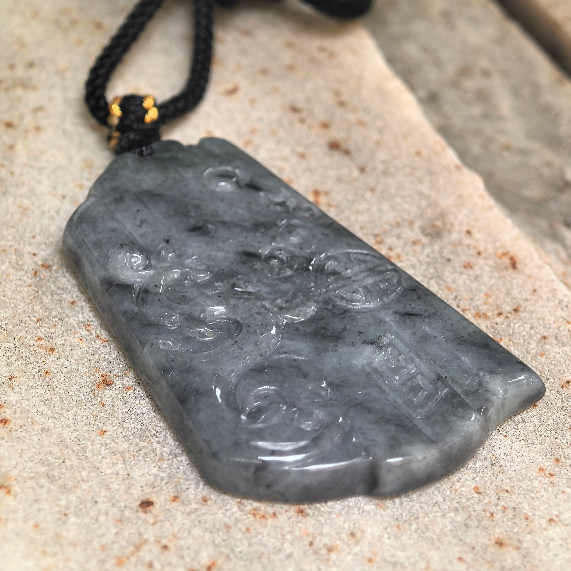Side view of gray-black carved jadeite pendant showing its slim plaque form and glossy luster.
