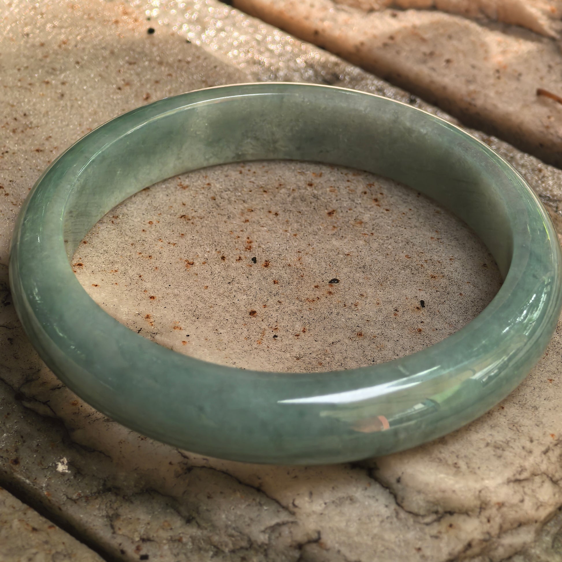 Natural Grade-A oil-green jadeite bangle displayed on stone surface with visible glutinous texture.
