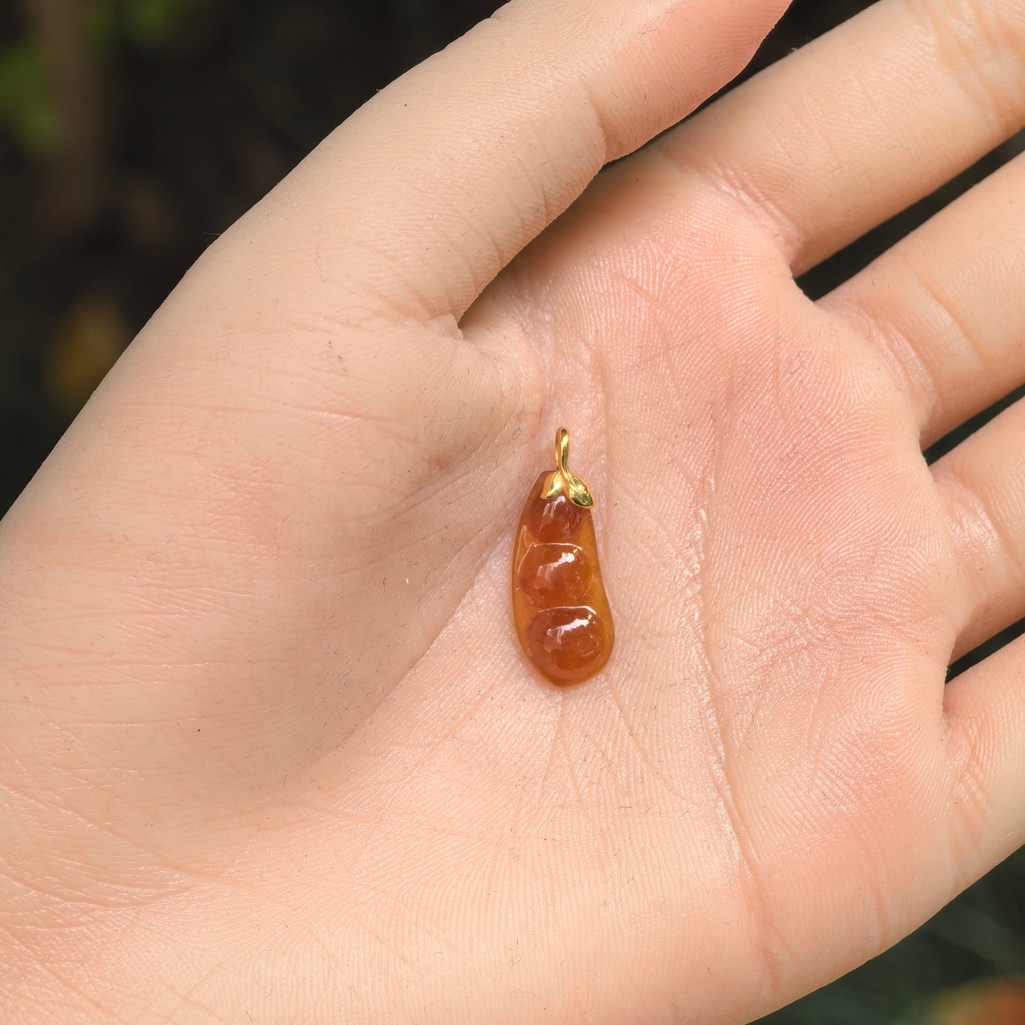 Small orange-red jadeite bean pendant with 18K gold leaf-style bail shown in hand.
