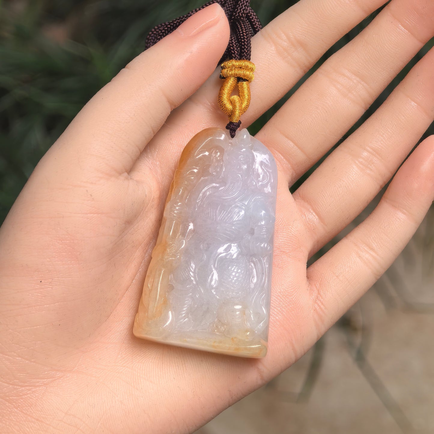 Natural Grade-A jadeite pendant on hand with Guan Gong carving and warm yellow-brown tone.

