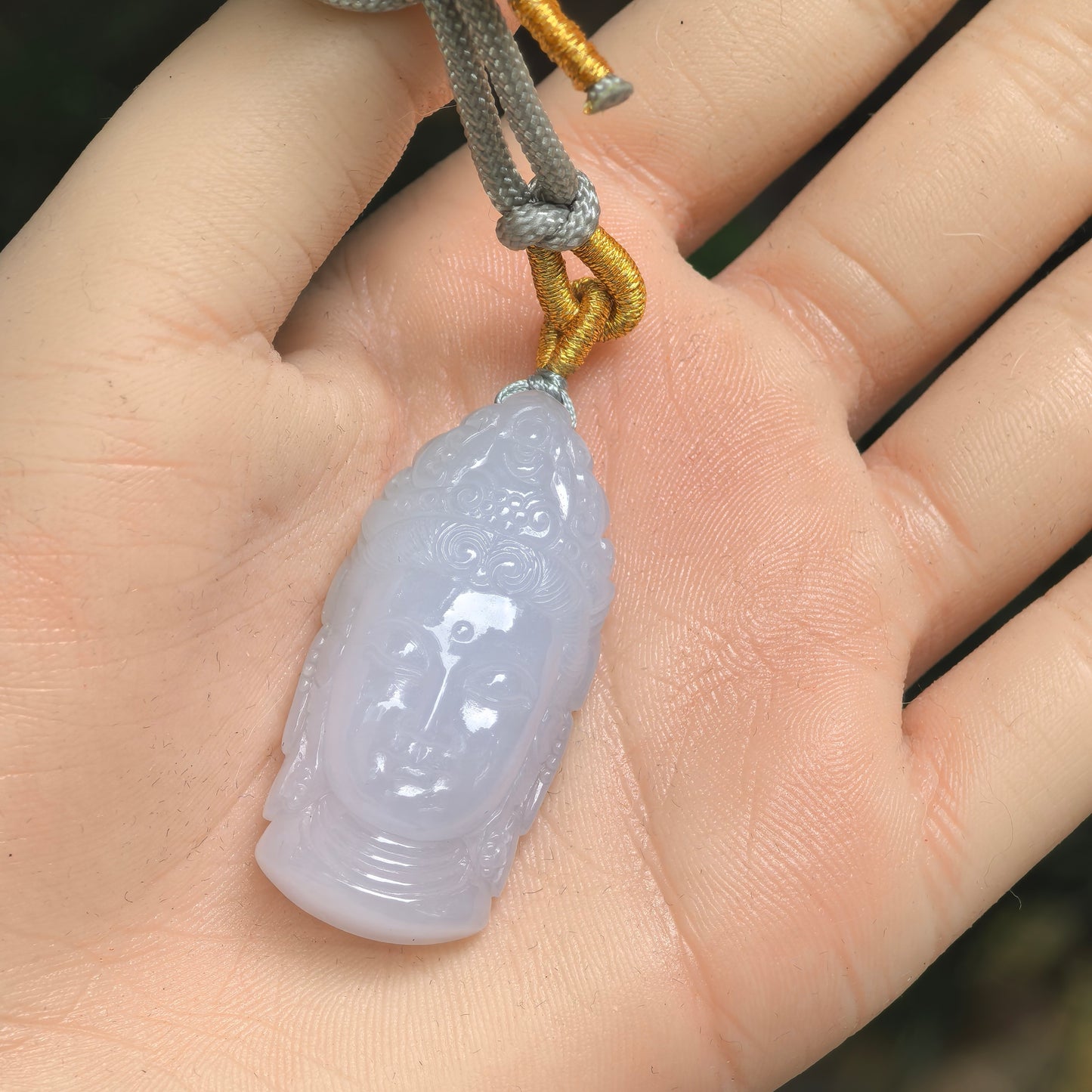 Pale lavender-white Burmese jadeite Buddha head pendant shown on hand with cord.
