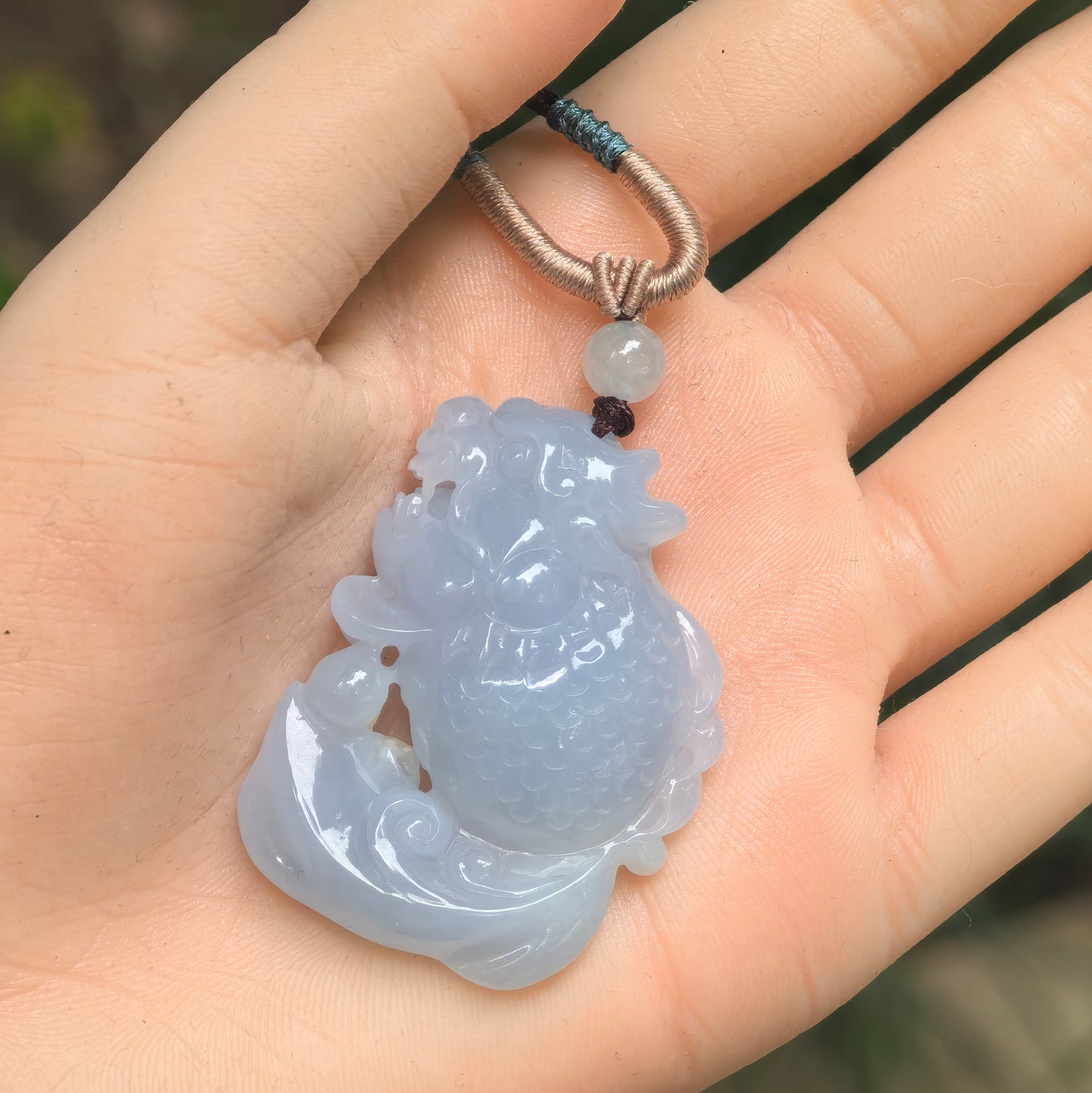 Pale lavender Burmese jadeite dragon-headed fish pendant shown on hand with cord.
