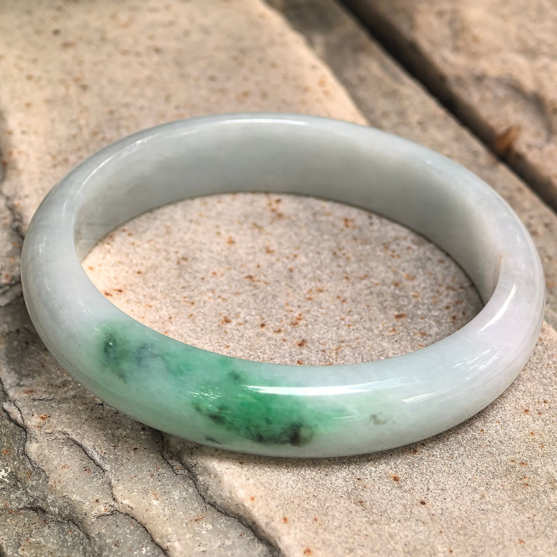 Glutinous green-white jadeite bangle placed on stone surface showing smooth rounded outer profile.
