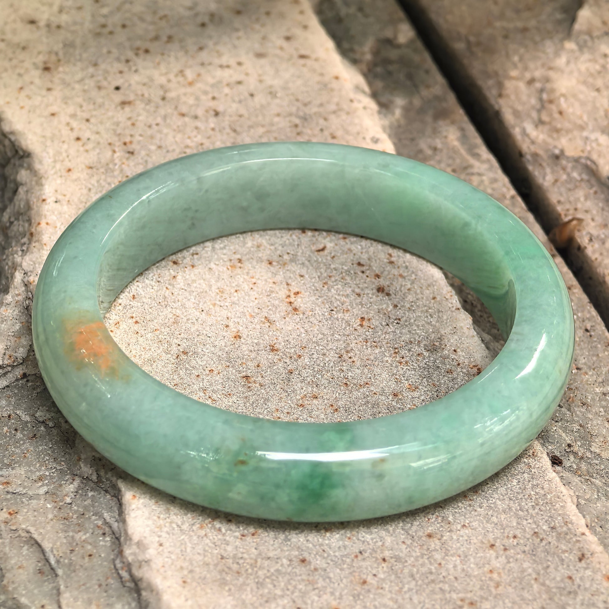 Glutinous green golden-flecked jadeite bangle placed on stone surface showing D-shape structure.
