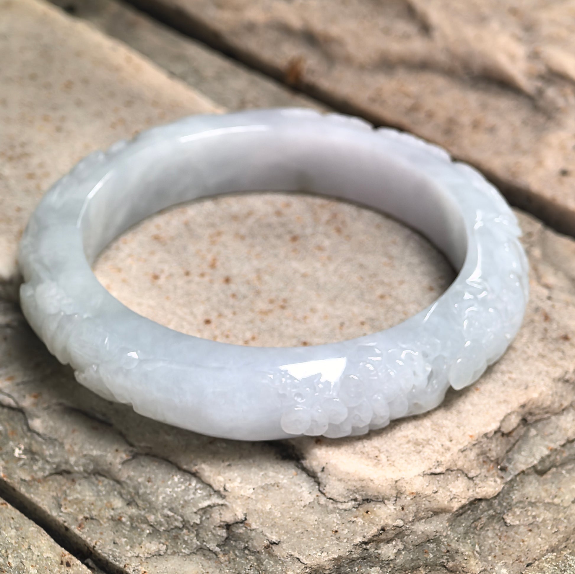 Natural Grade-A white jadeite bangle displayed on stone background with carved floral design.
