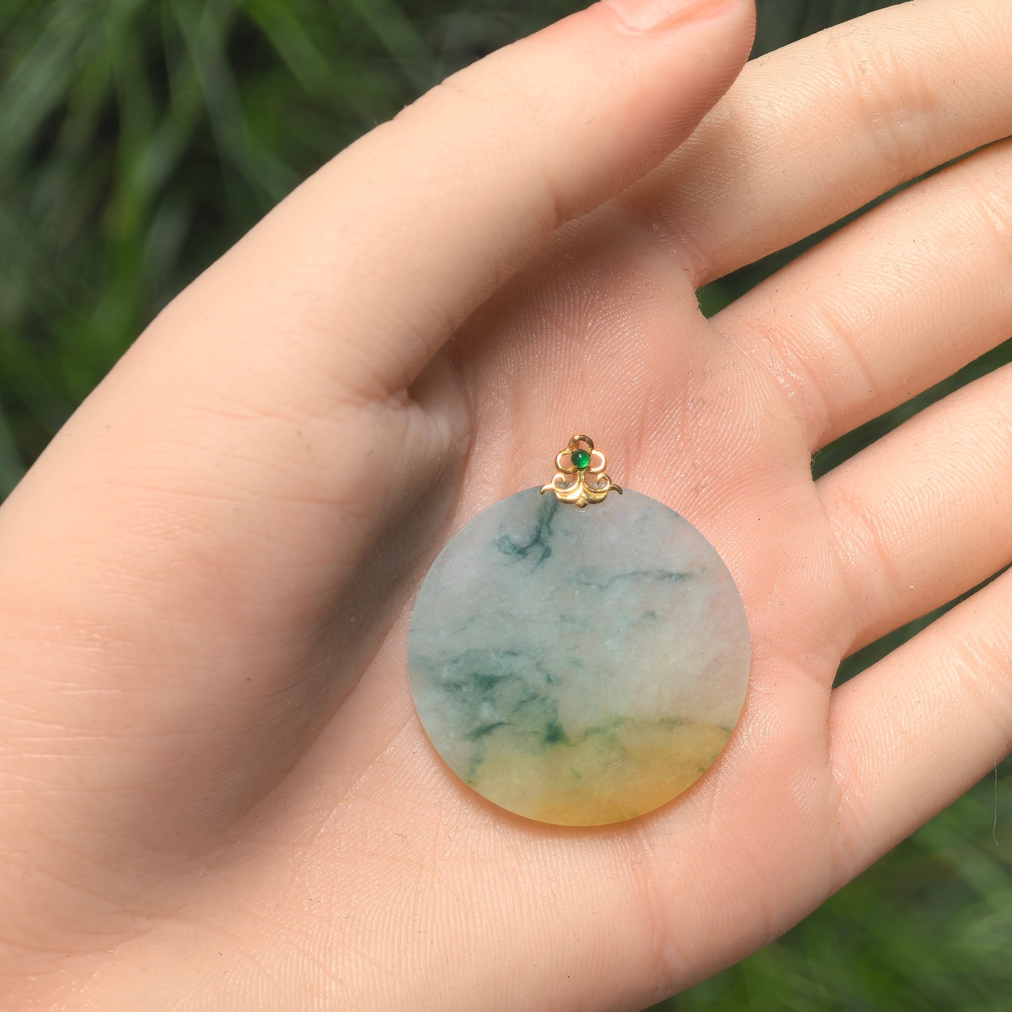 Natural Grade-A Burmese jadeite pendant held in hand showing round shape and 18K gold bail.
