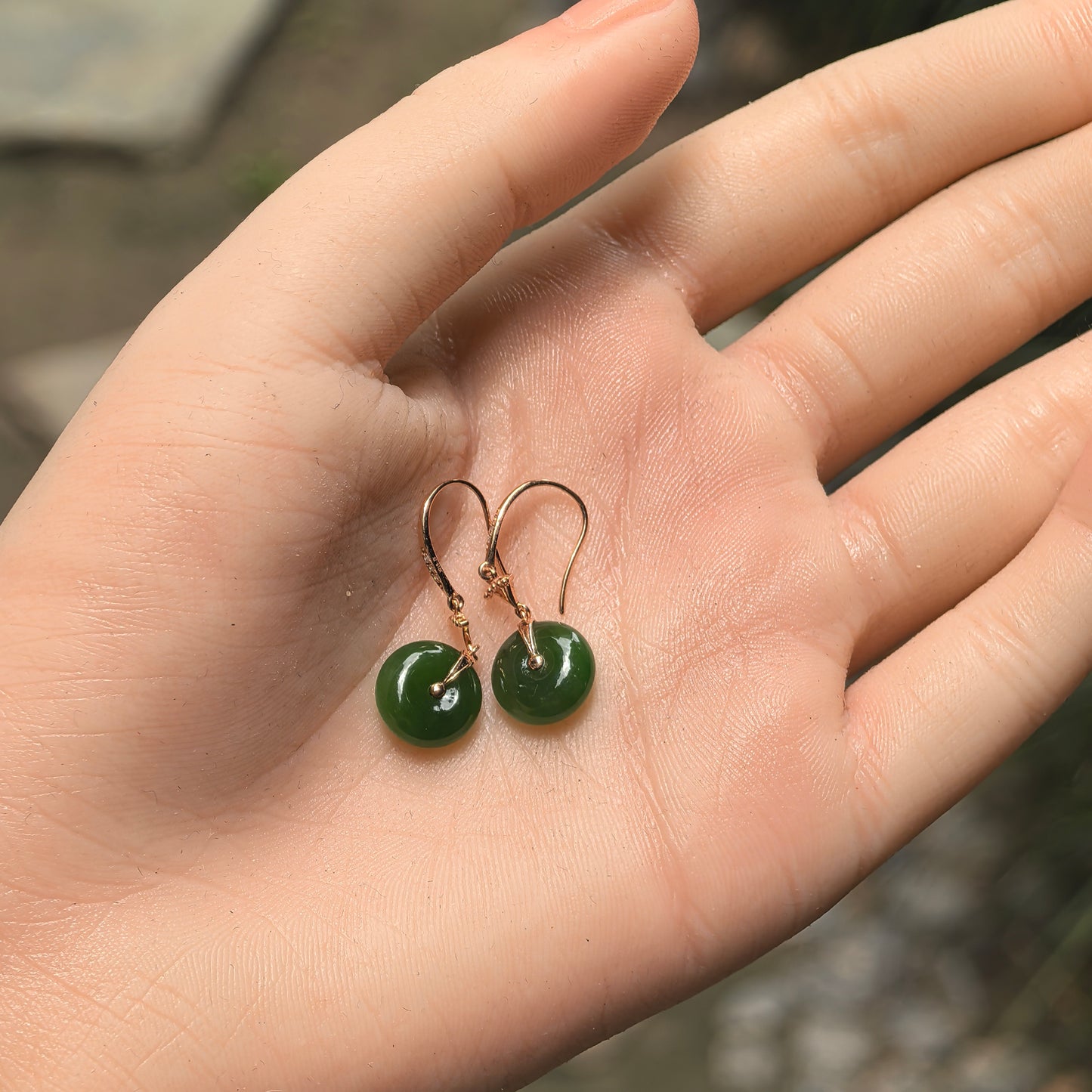 Hand-held pair of natural deep green Hetian jade donut earrings with polished 18K gold hooks and connectors.