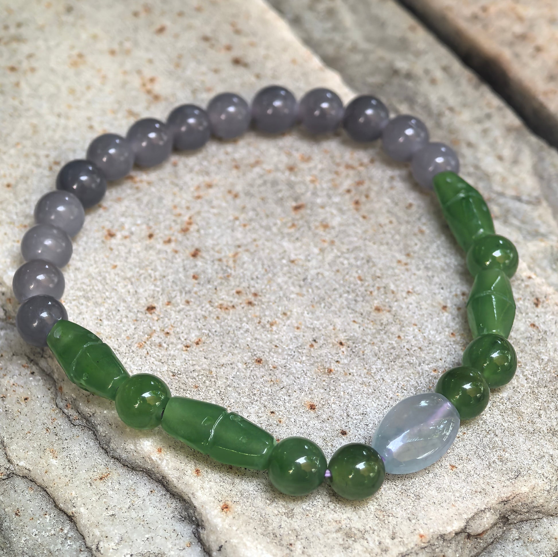 Close-up of the natural green and smoky lavender jade bracelet arranged on a textured stone surface.