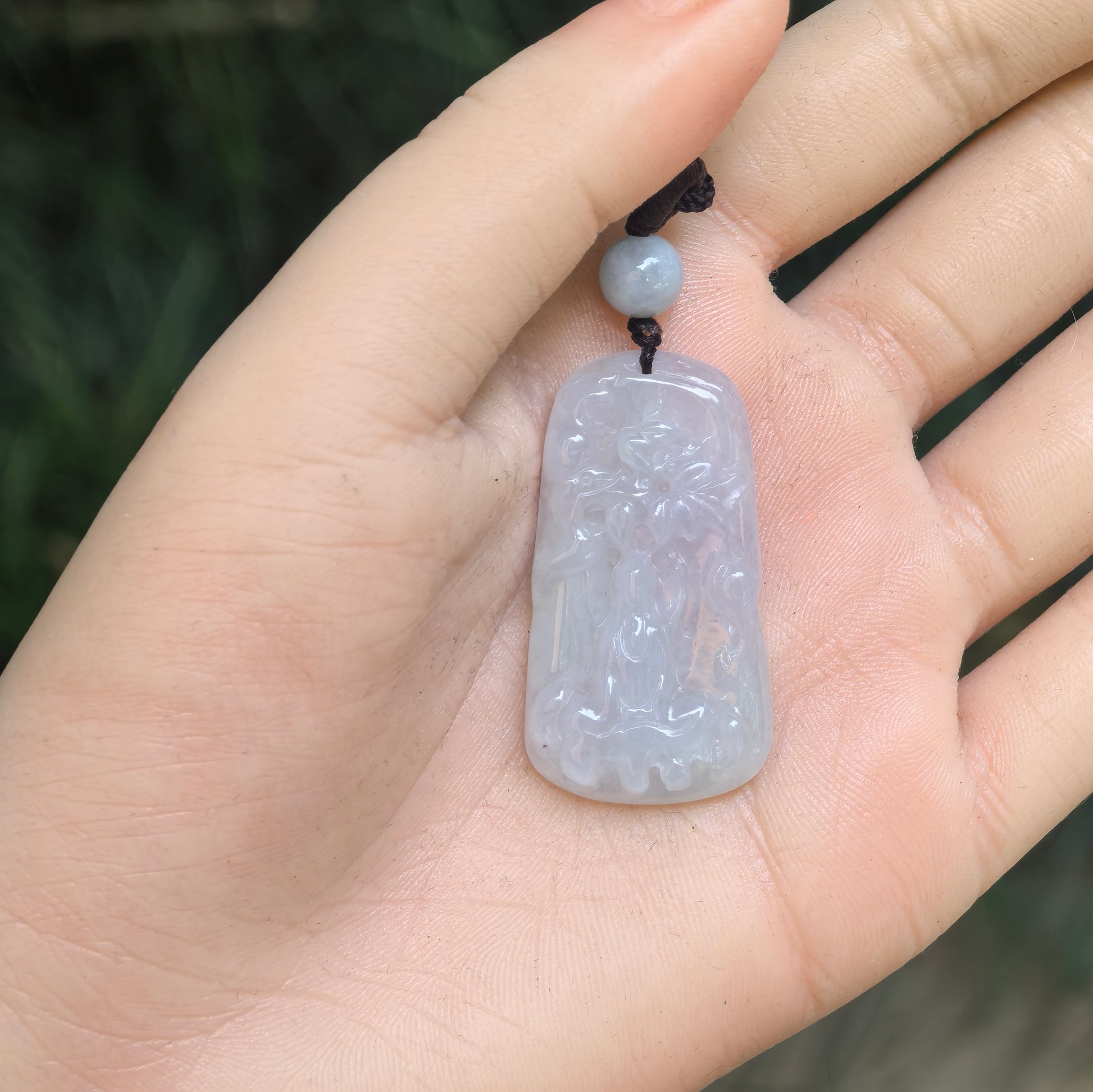 Hand-held view showing the size, rectangular shape, decorative bead, and woven cord of the white jadeite pendant.