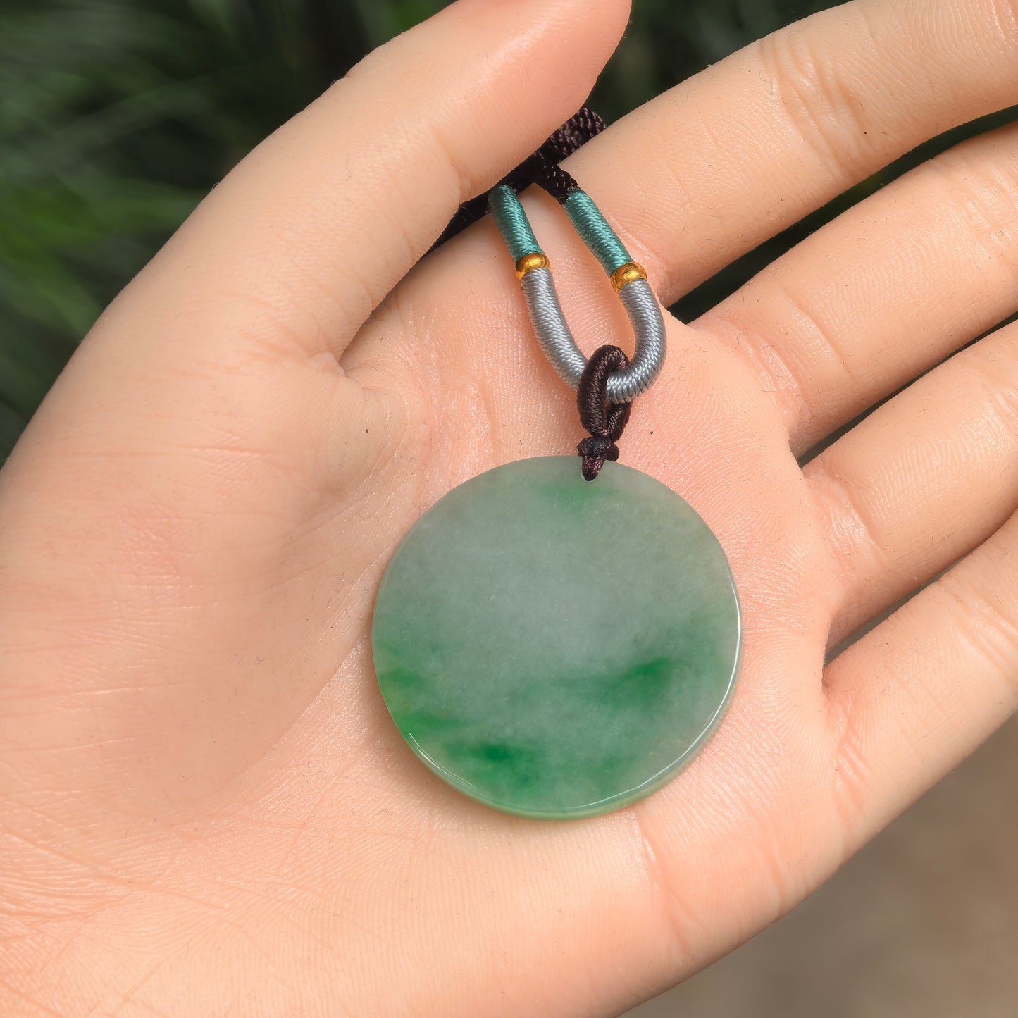 Hand-held view of a round green floral Burmese jadeite plain plaque pendant showing its size and natural color distribution.