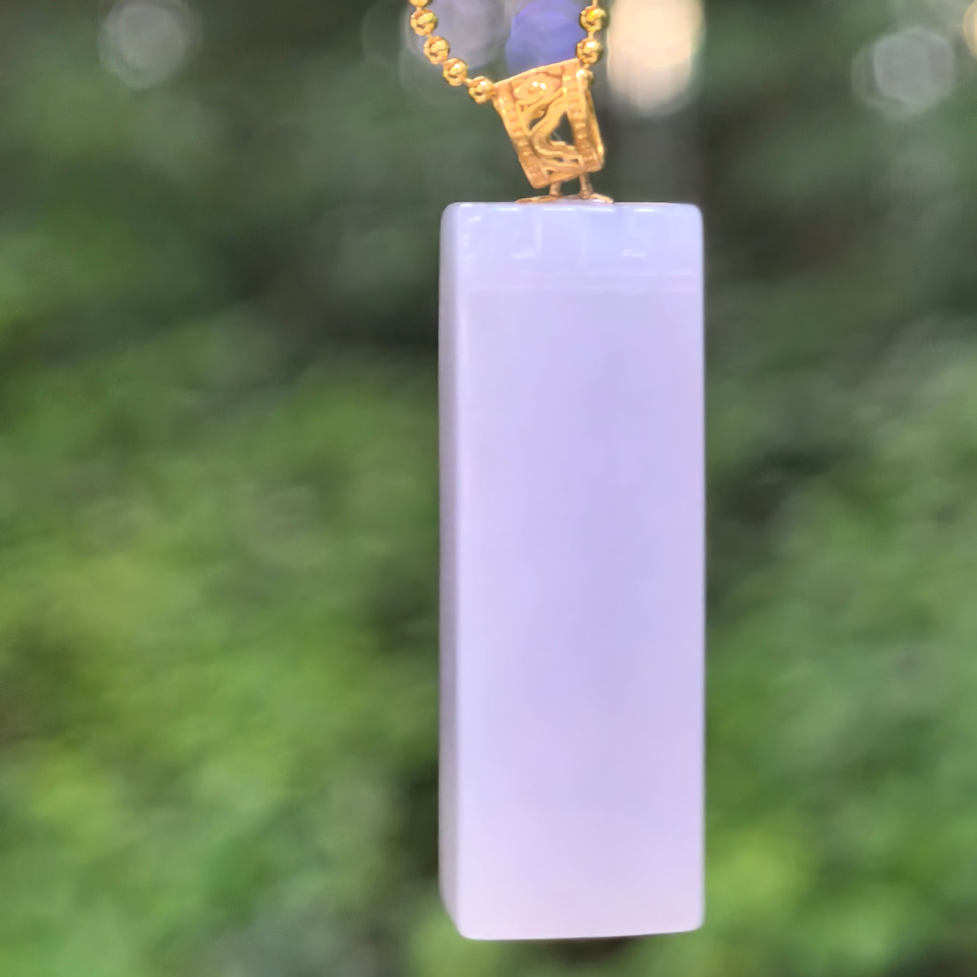 Natural Grade-A icy-glutinous pale lavender jadeite column pendant with an 18K gold bail hanging in natural light.