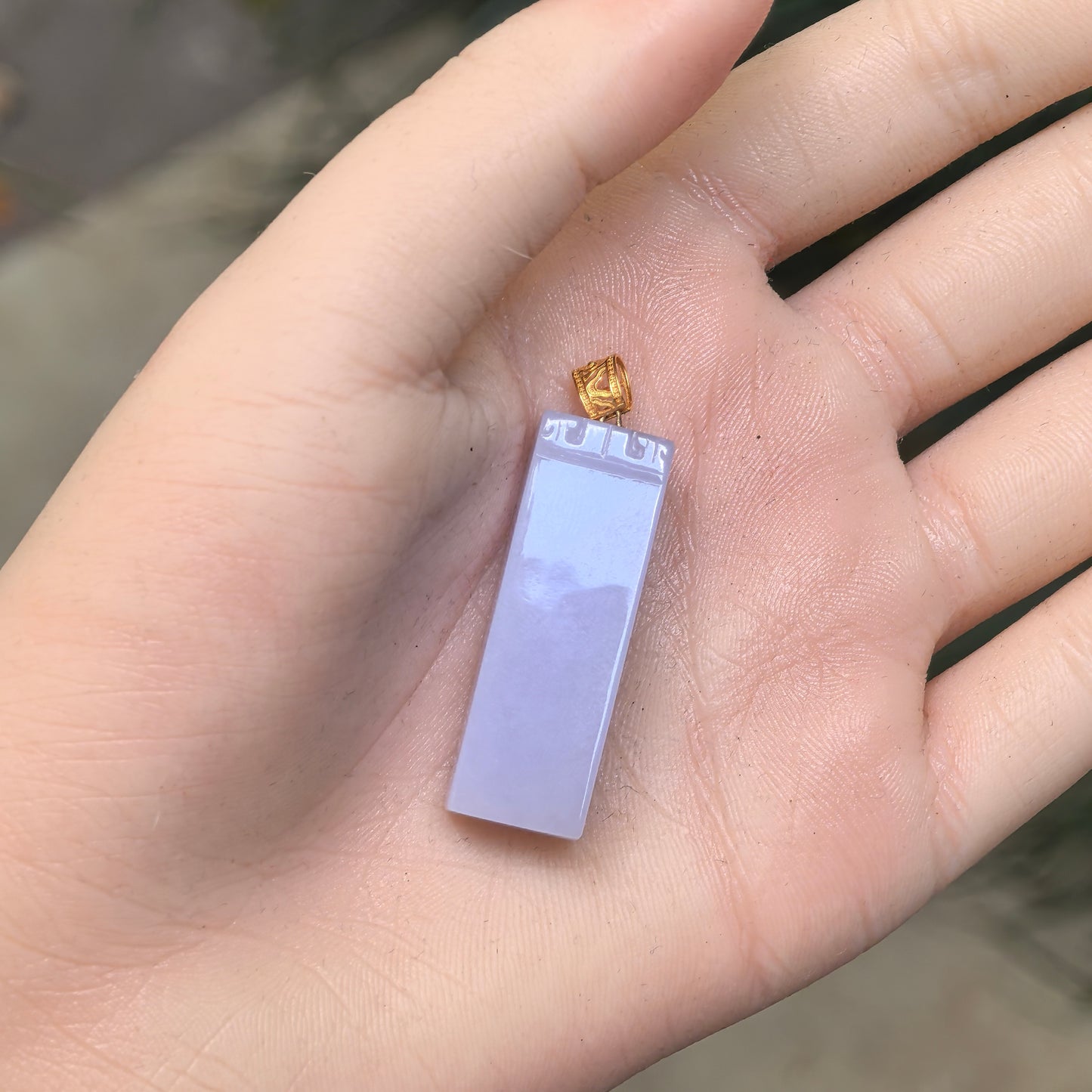 Hand-held view of the pale lavender jadeite pendant showing its elongated rectangular proportions and polished plain surface.