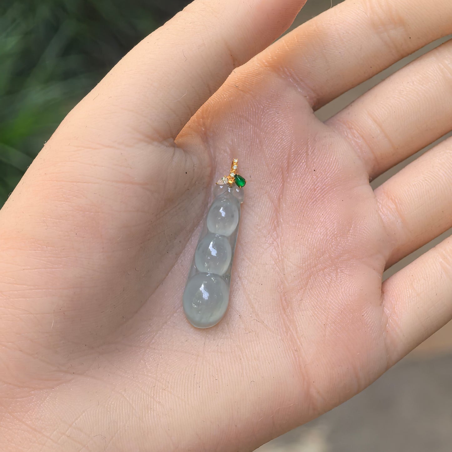 Hand-held view of the light green three-bean jadeite pendant showing its petite elongated proportions and translucent texture.