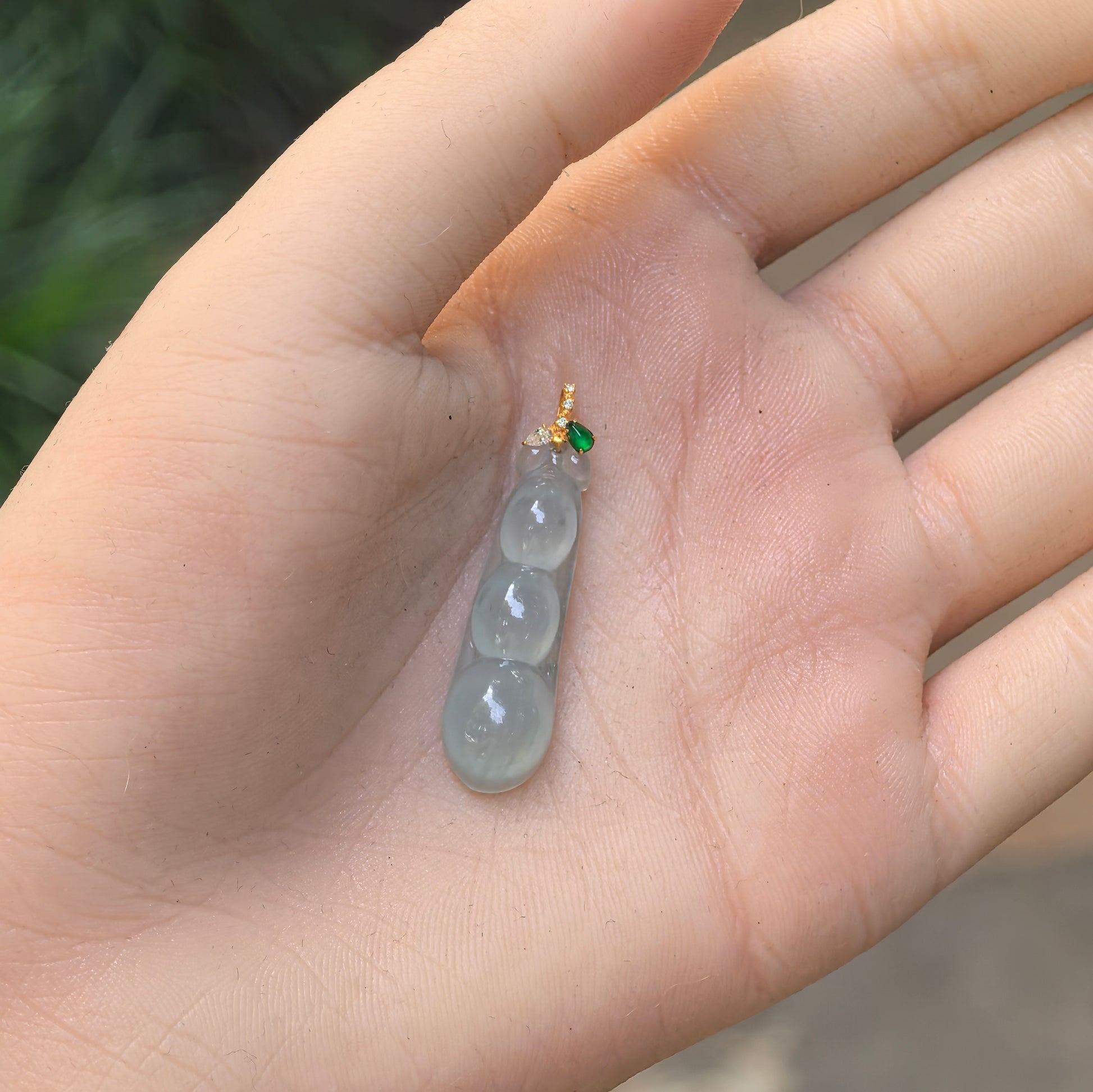 Hand-held view of the light green three-bean jadeite pendant showing its petite elongated proportions and translucent texture.