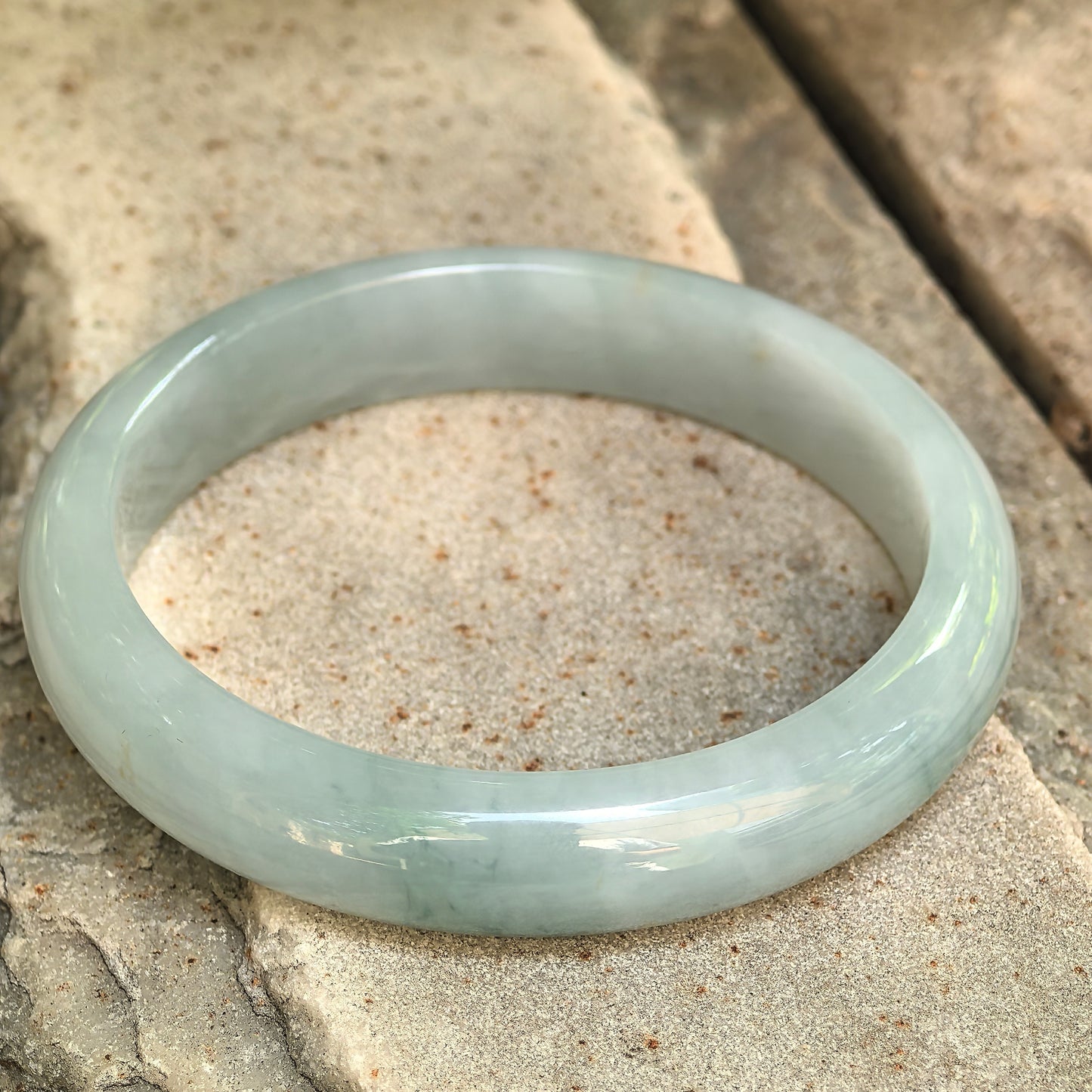 Natural Grade-A light green jadeite D-shape bangle displayed on stone under natural light.