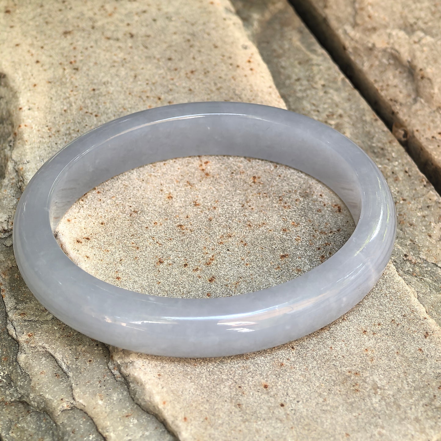 Natural Grade-A blue-lavender jadeite oval bangle displayed on stone, showing its smooth rounded profile.