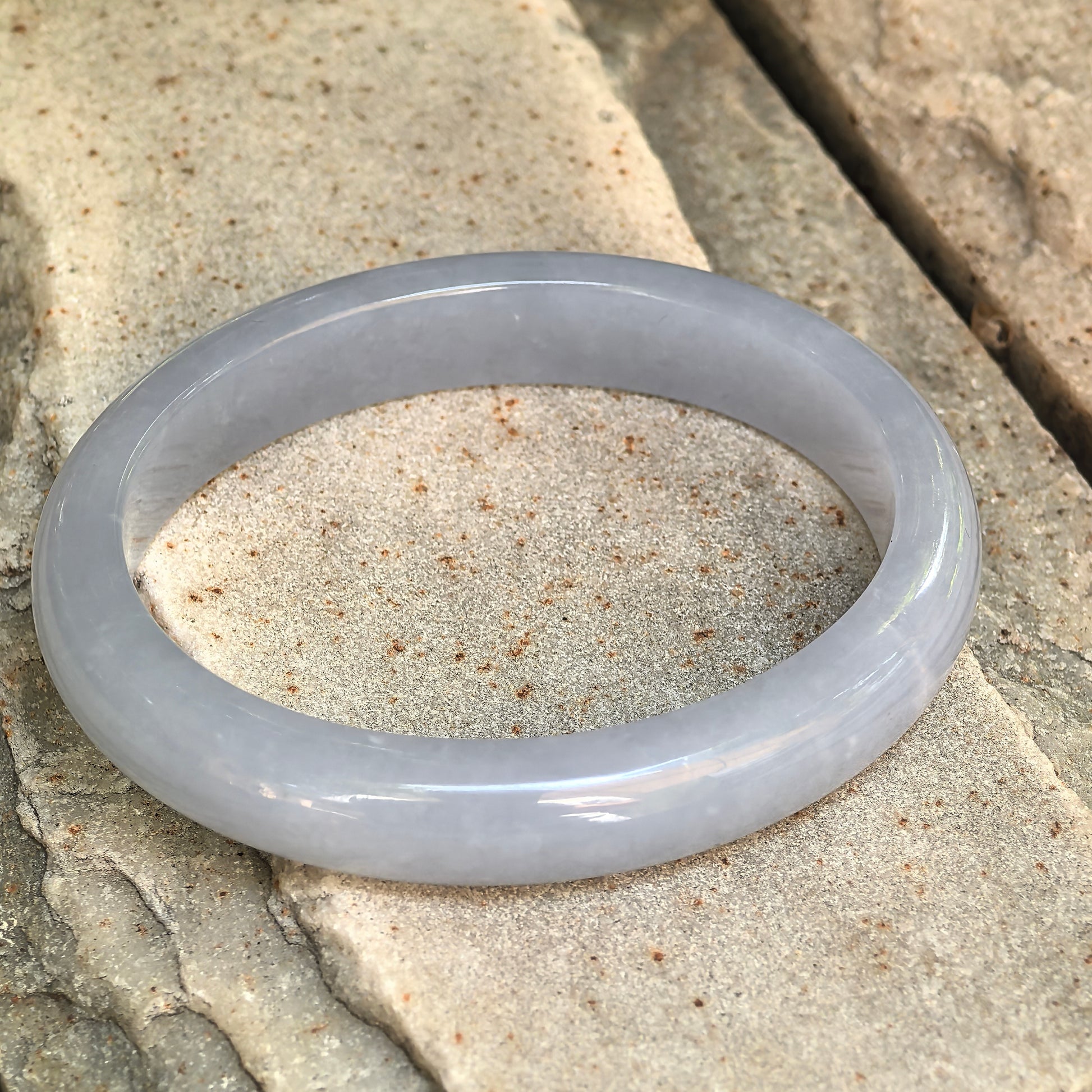 Natural Grade-A blue-lavender jadeite oval bangle displayed on stone, showing its smooth rounded profile.