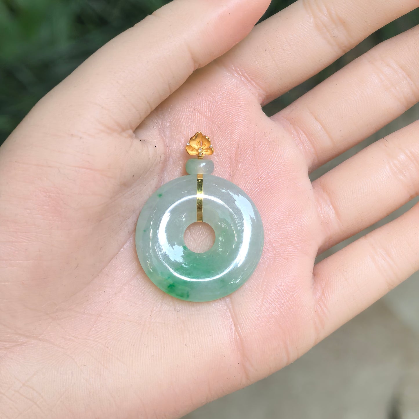 Light green Burmese jadeite donut pendant shown in the hand to display its size, rounded profile, and 18K gold setting.