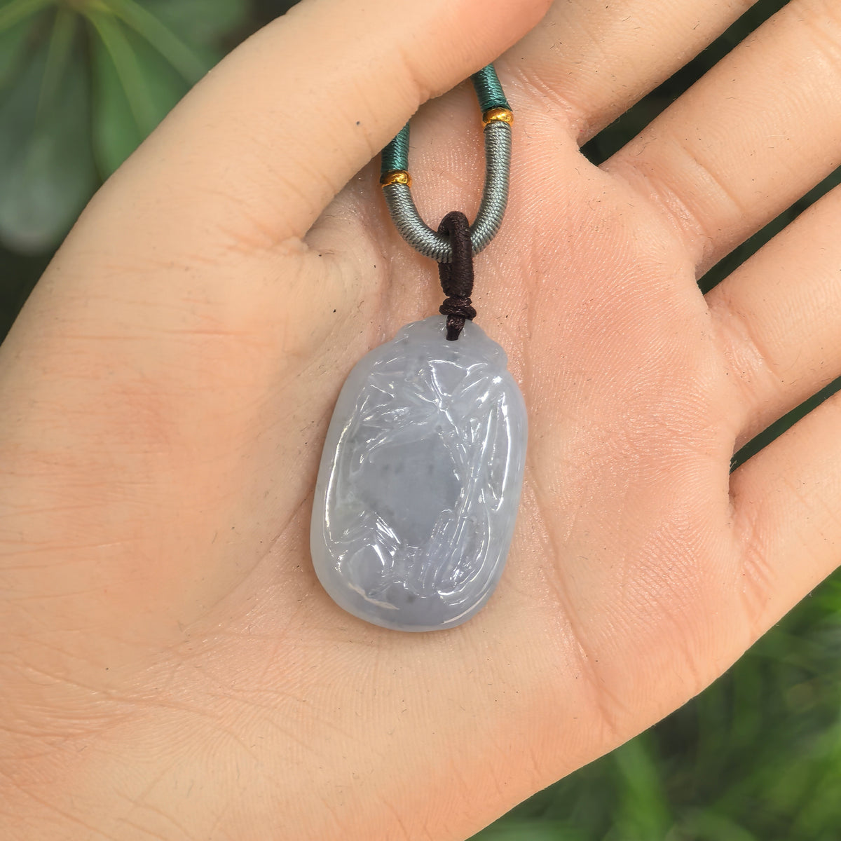 Hand-held view of the pale lavender Burmese jadeite pendant showing its bamboo carving and rounded rectangular shape.