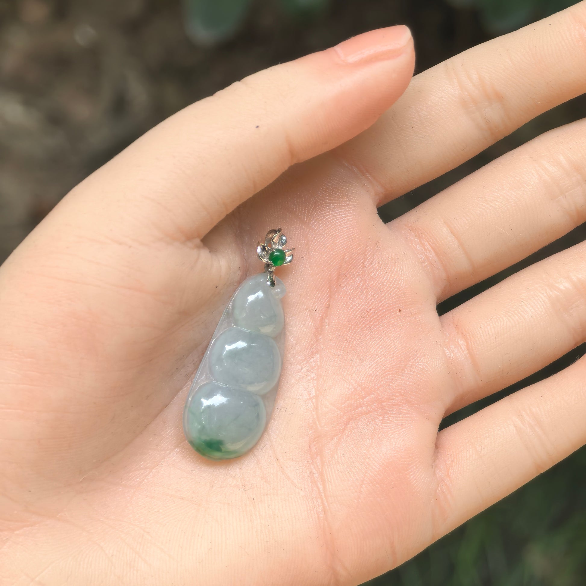 Hand-held view of a slender light green three-bean jadeite pendant with a deeper green accent.