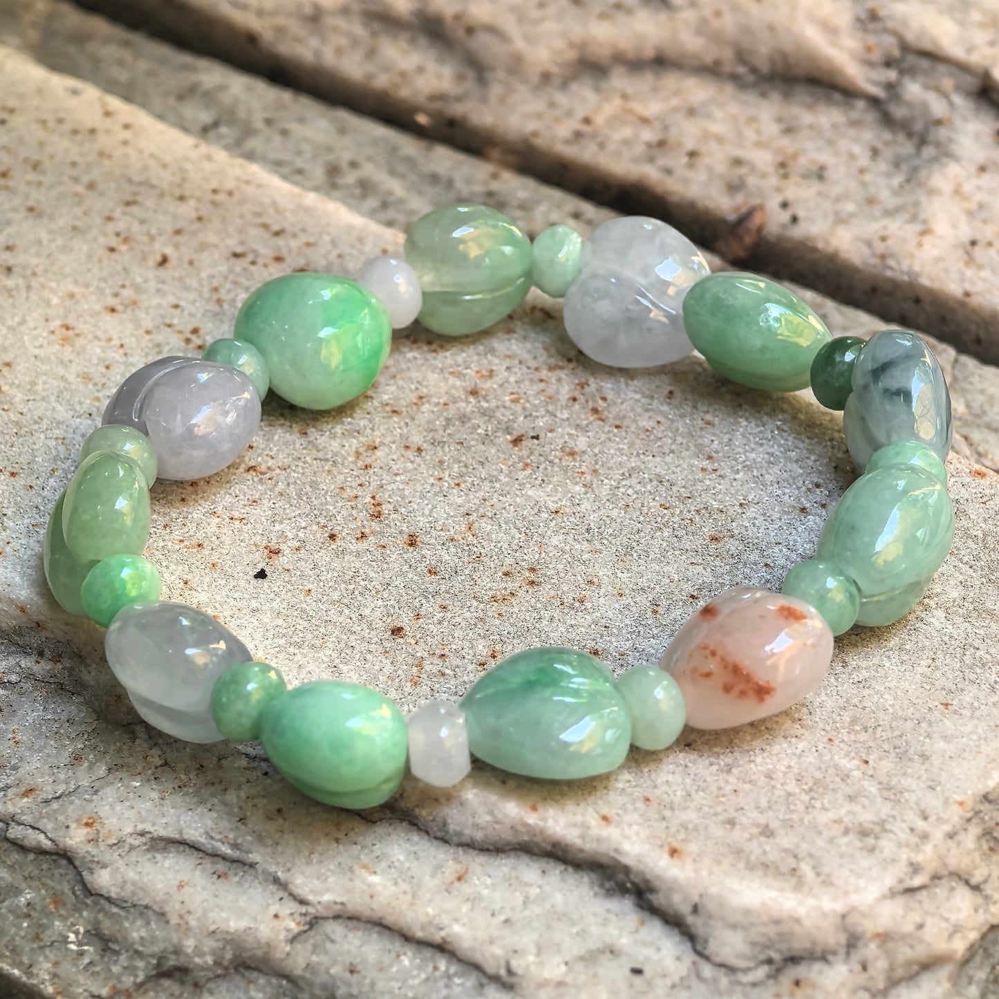 Natural Grade-A green and lavender peach-shaped jadeite bracelet arranged on a textured stone surface.