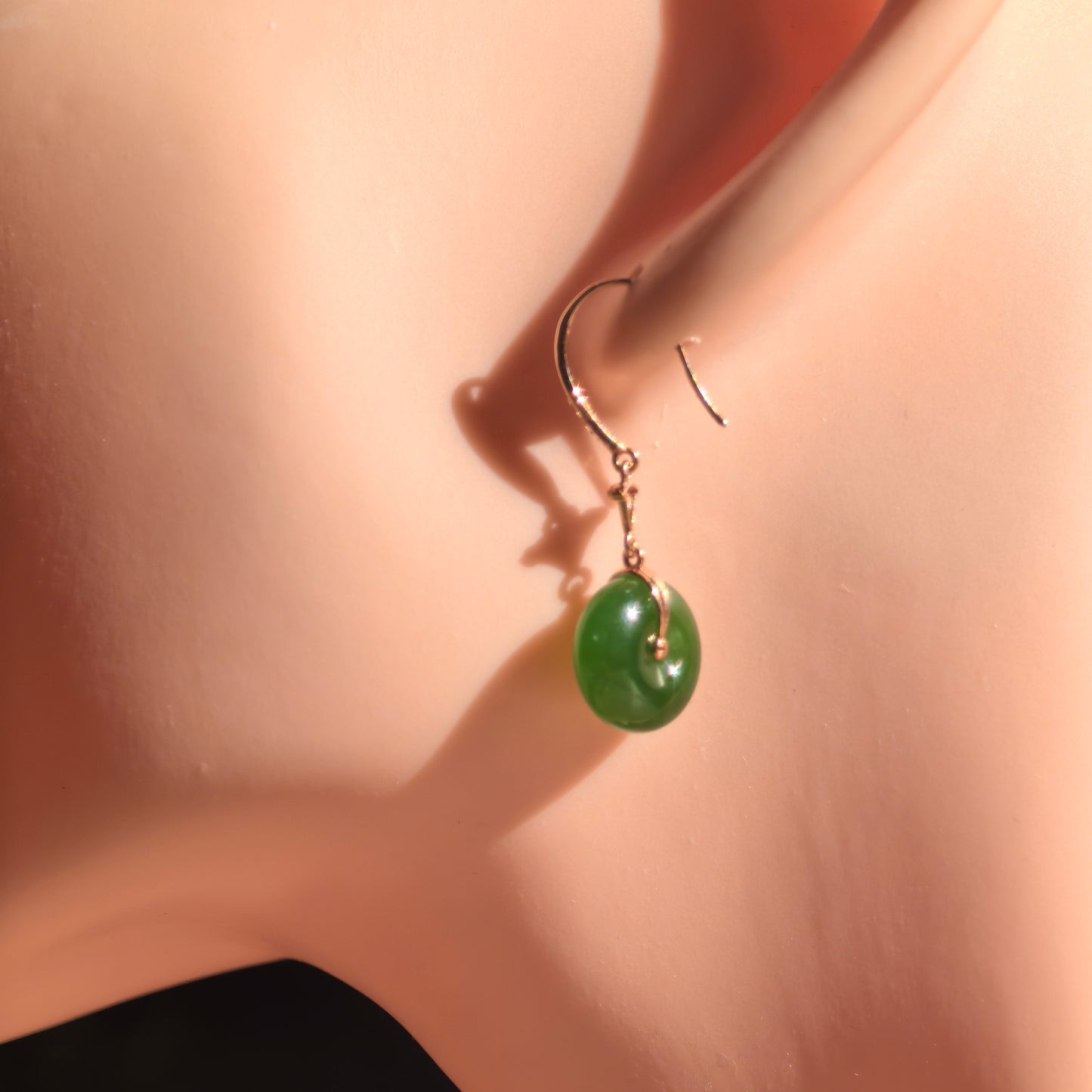 Deep green Hetian jade drop earring worn on an ear display, showing its rounded donut shape and glossy oily luster.