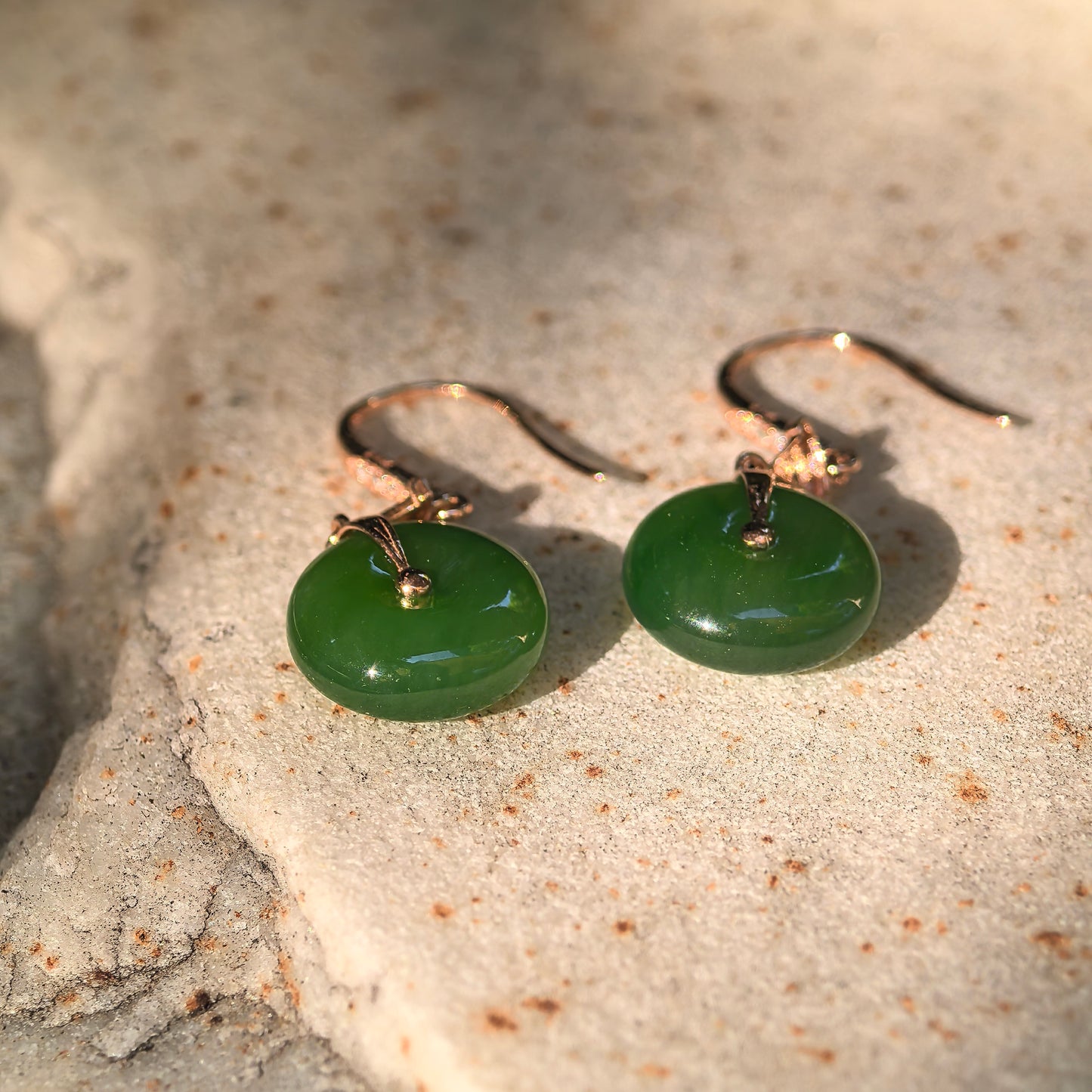 Pair of deep green Hetian jade donut earrings displayed on a textured stone surface under warm sunlight.