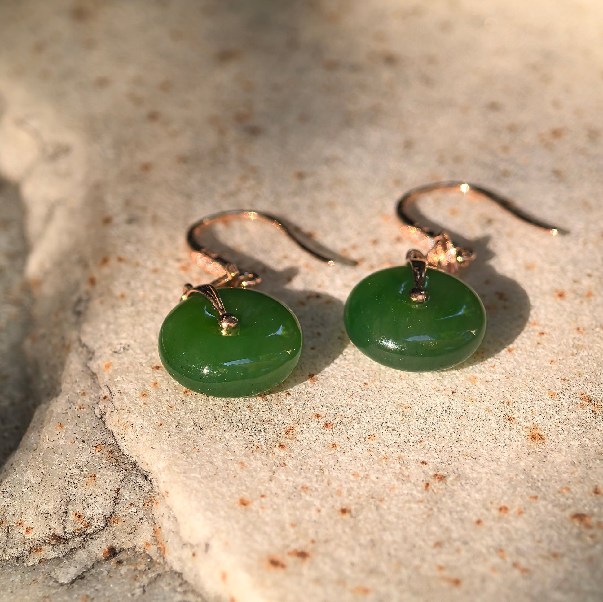 Pair of deep green Hetian jade donut earrings displayed on a textured stone surface under warm sunlight.