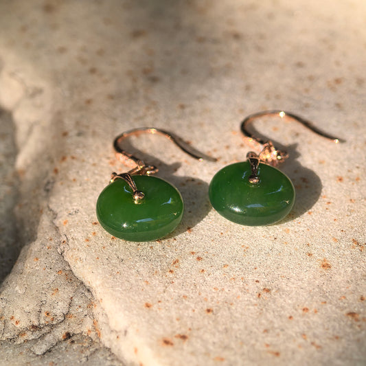 Pair of deep green Hetian jade donut earrings displayed on a textured stone surface under warm sunlight.