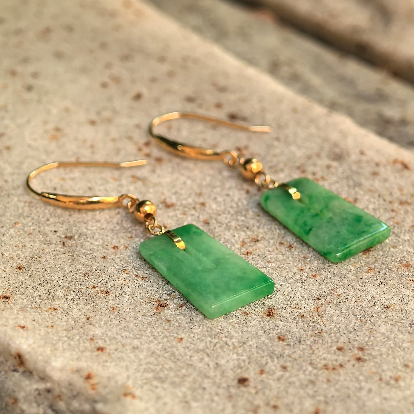 Pair of glutinous green jadeite earrings with 18K gold hooks displayed on a textured stone surface.
