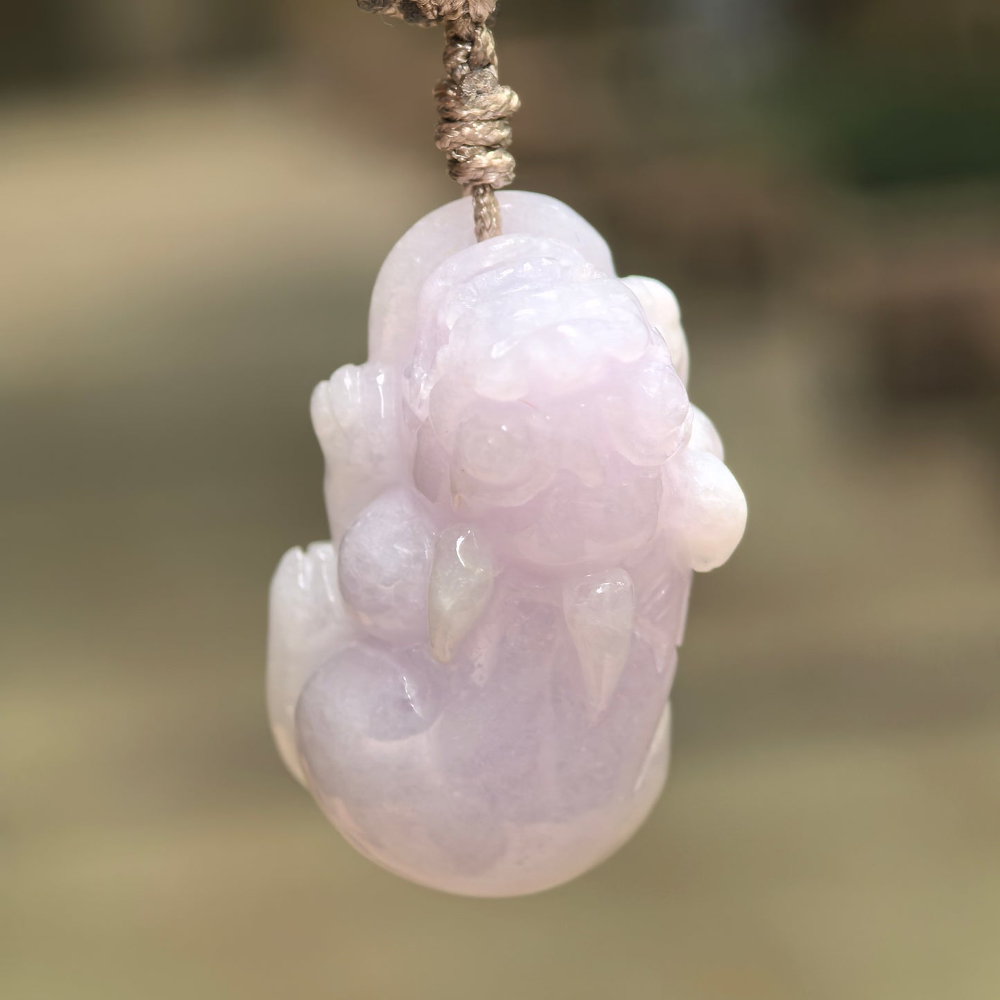Natural Grade-A pale lavender Pixiu jadeite pendant hanging from its braided cord against a softly blurred background.