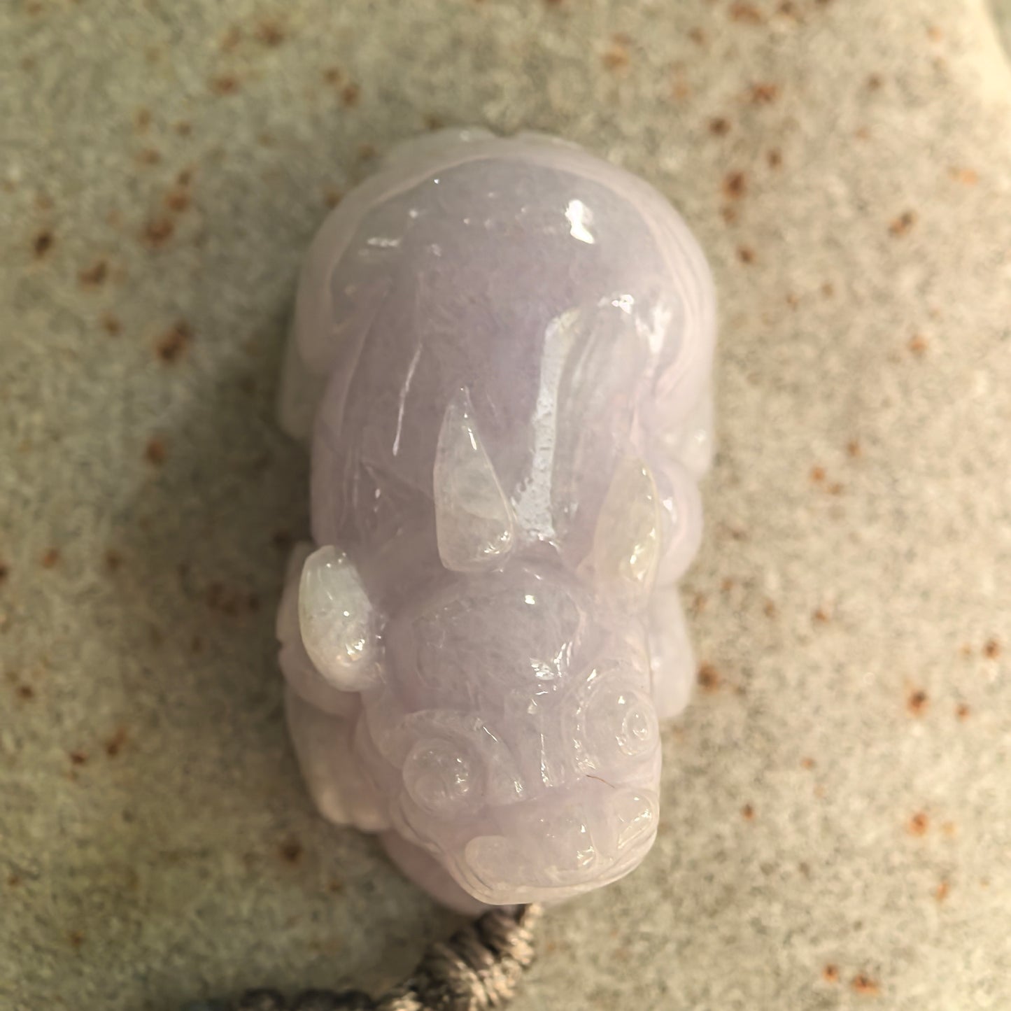 Detailed front view of a glutinous pale lavender Burmese jadeite Pixiu pendant resting on a stone surface.