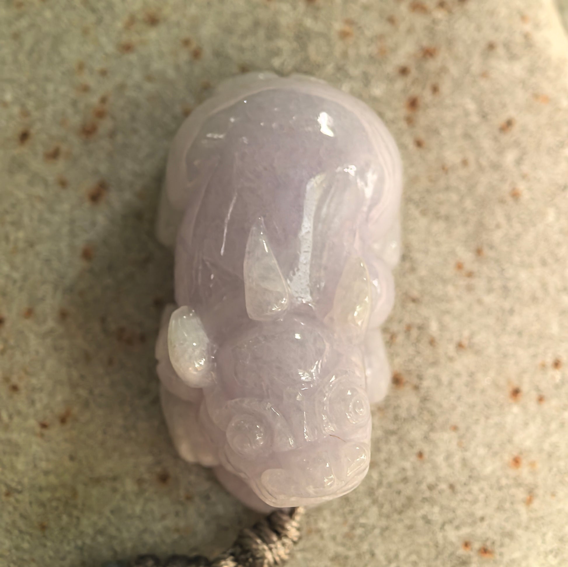Detailed front view of a glutinous pale lavender Burmese jadeite Pixiu pendant resting on a stone surface.