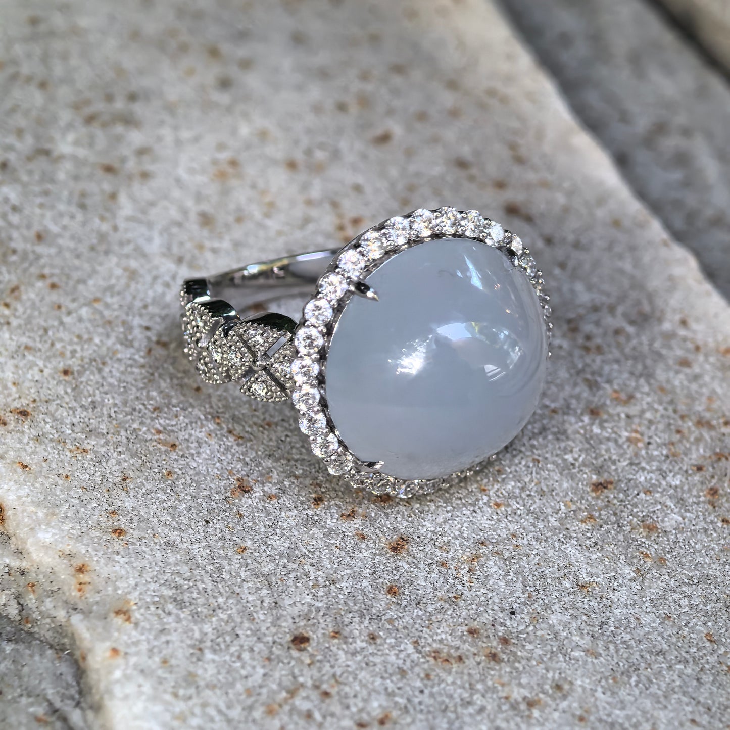 Natural Grade-A icy glutinous light blue cabochon jadeite ring in Pt950 platinum with synthetic diamond halo displayed on stone.