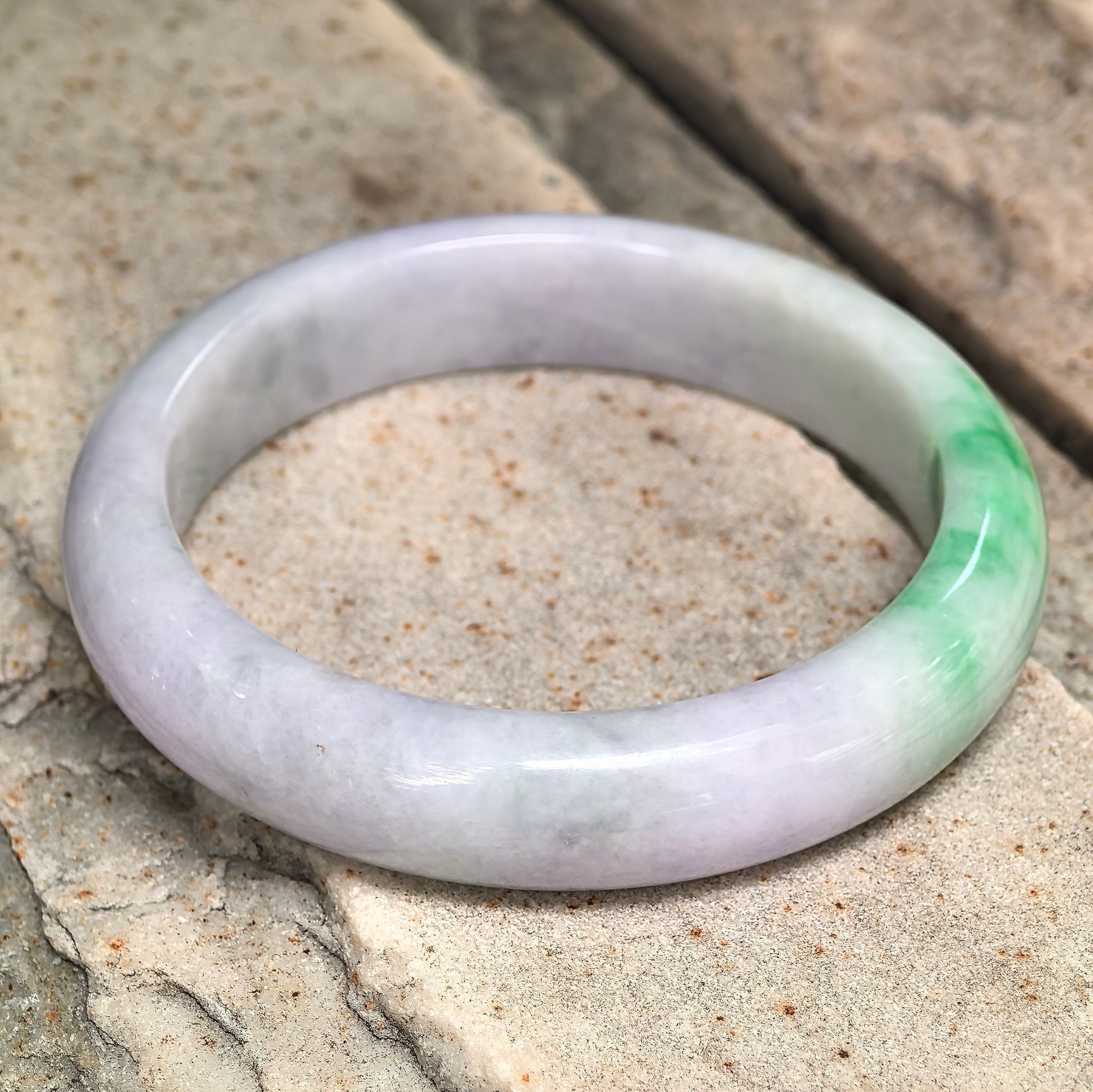 Natural Grade-A lavender-green D-shape jadeite bangle displayed on stone, showing the pale lavender base and bright green section.